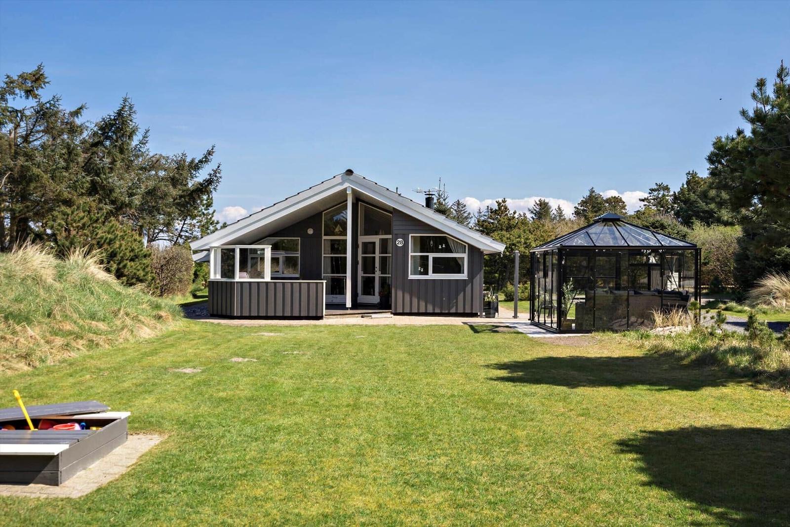 Moderne Holzhütte mit Garten, Terrasse und Glasüberdachung. Umgeben von Gras und Bäumen.