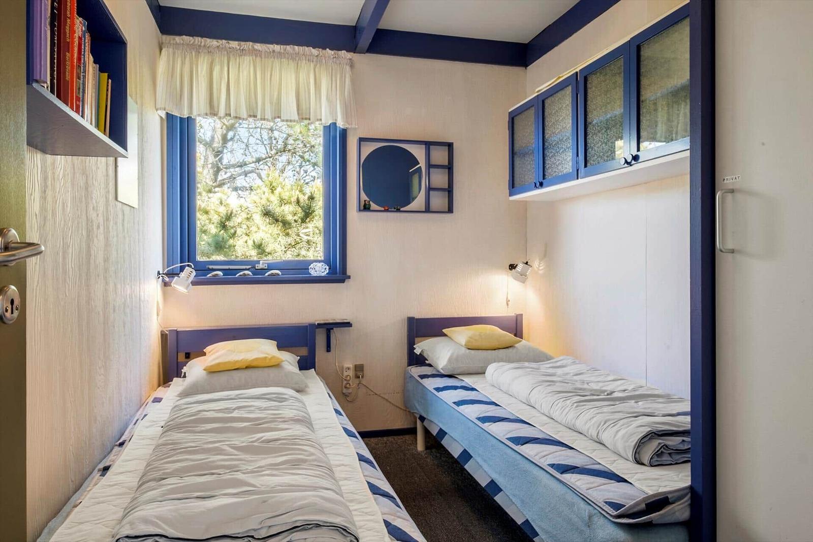 Zwei Einzelbetten mit blauen Kopfenden und gelben Kissen. Fenster mit blauem Rahmen und Bücherregal.