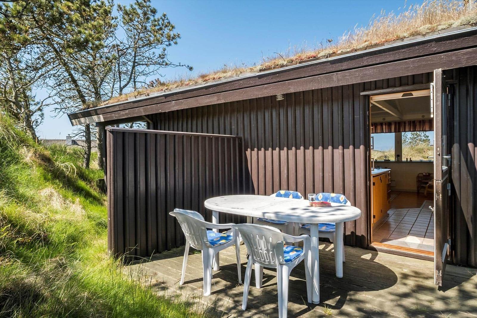 Hütte mit Grasdach und Terrasse mit Tisch und Stühlen. Küche sichtbar durch offene Tür.