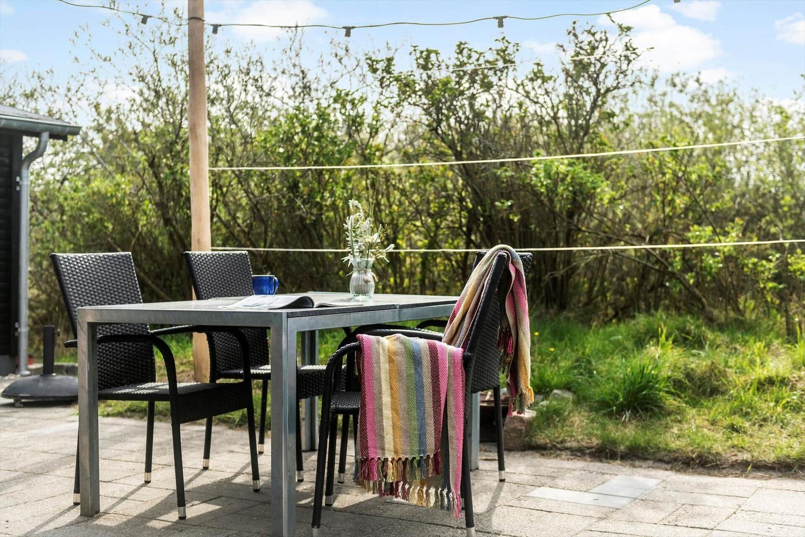 Terrasse mit Tisch, vier Stühlen und Decke. Hintergrund: Grünflächen und Bäume.