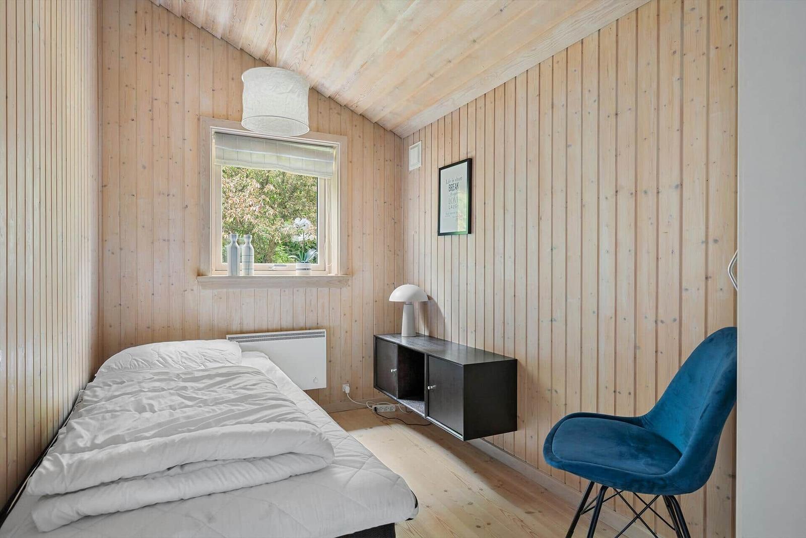 Schlafzimmer mit Holzwand, Bett, blauer Sessel und Fenster mit Blick auf Garten.
