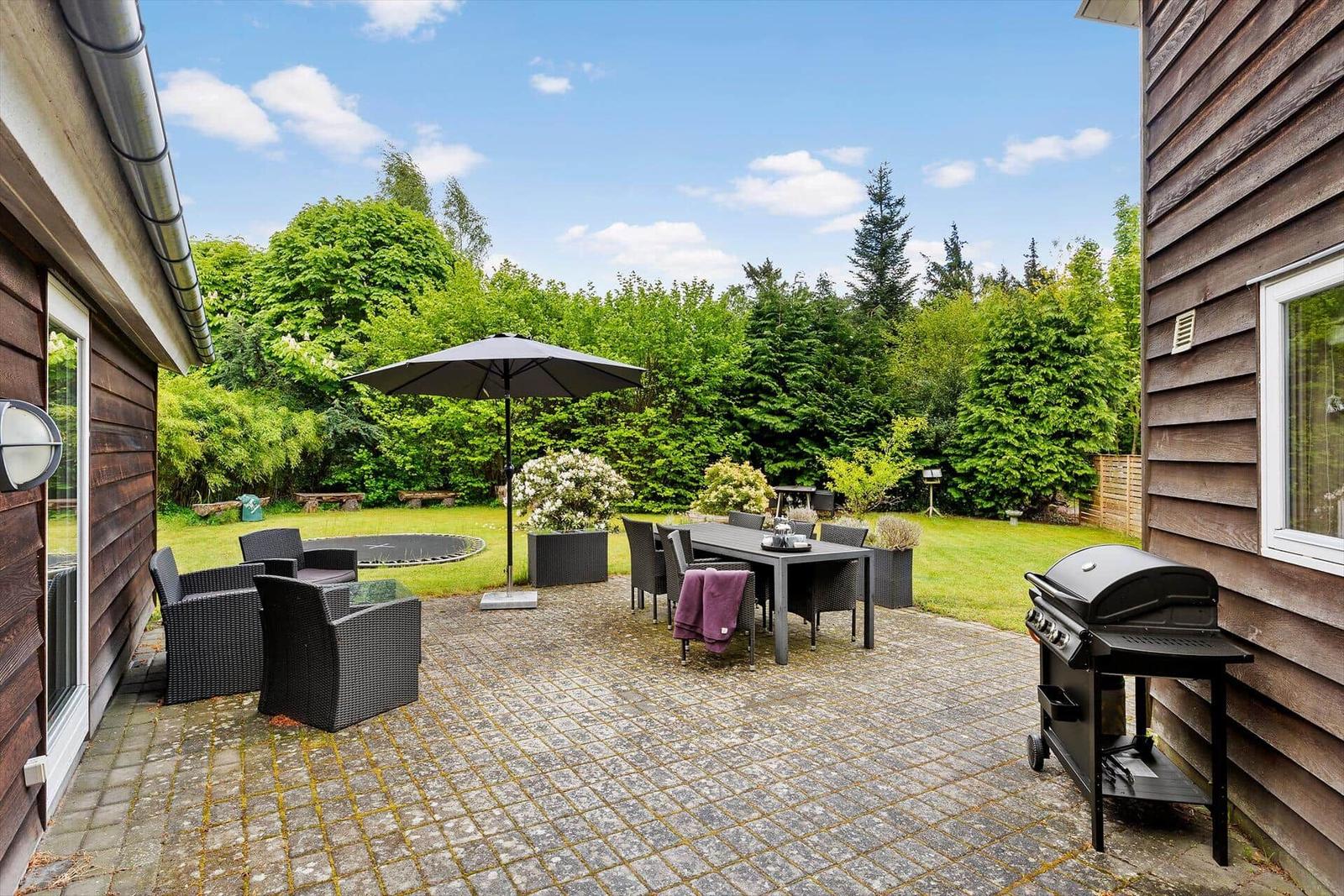 Terrasse mit Grill, Tisch, Stühlen und Sonnenschirm. Grünflächen und Bäume im Hintergrund.