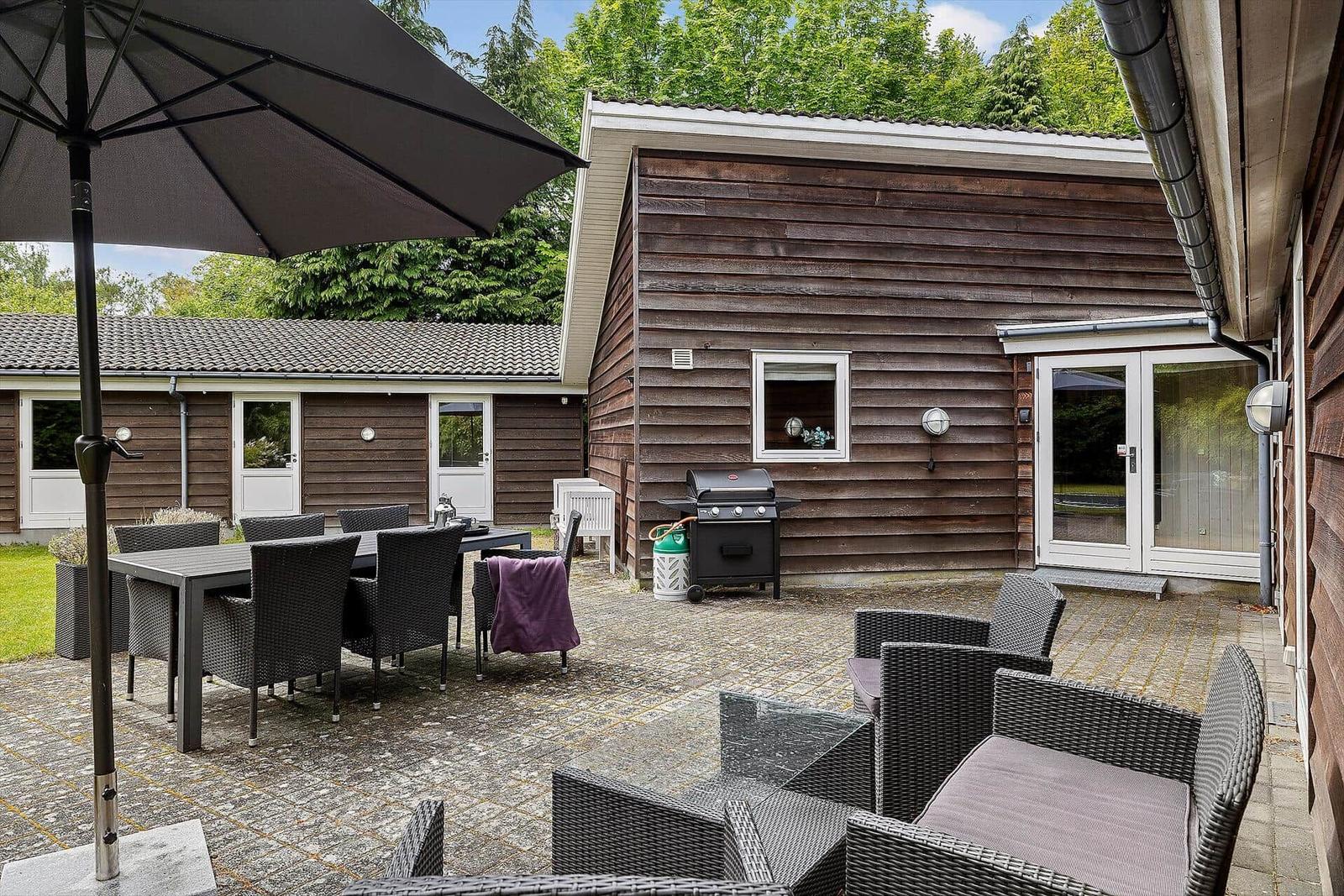 Terrasse mit Sitzgruppe, Tisch, Sonnenschirm und Grill. Holzhaus mit Fenster und Glastür.