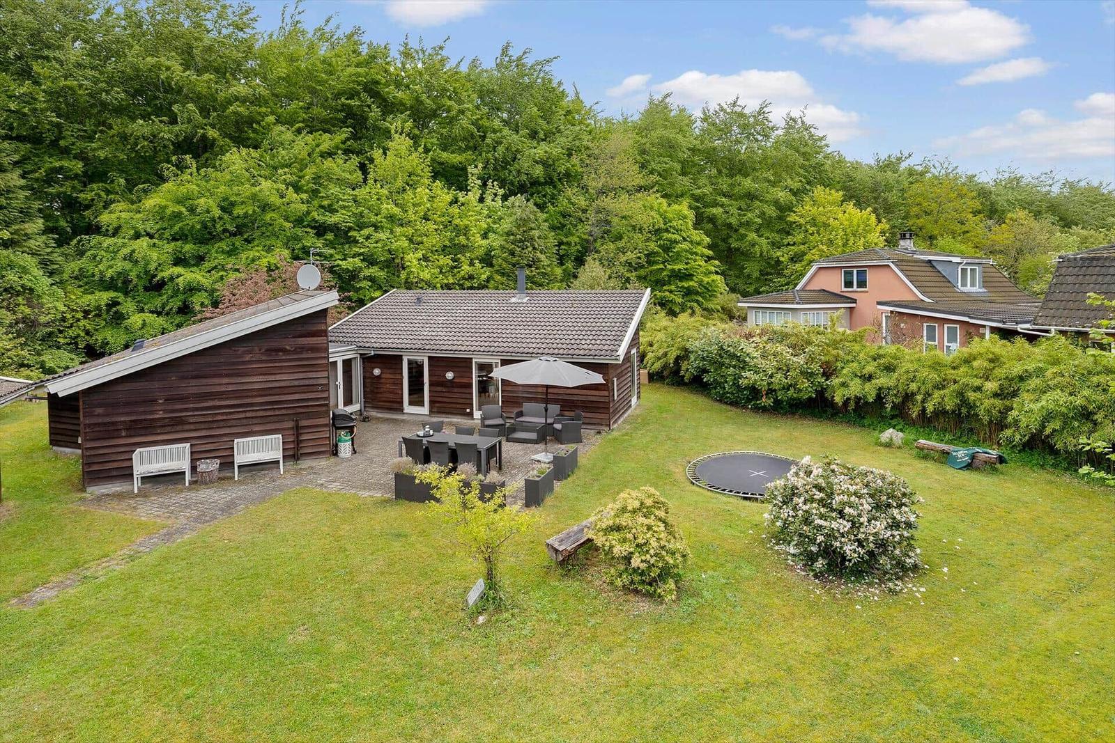 Holzhaus mit Terrasse, Garten und Trampolin. Umgeben von Bäumen und Grün.