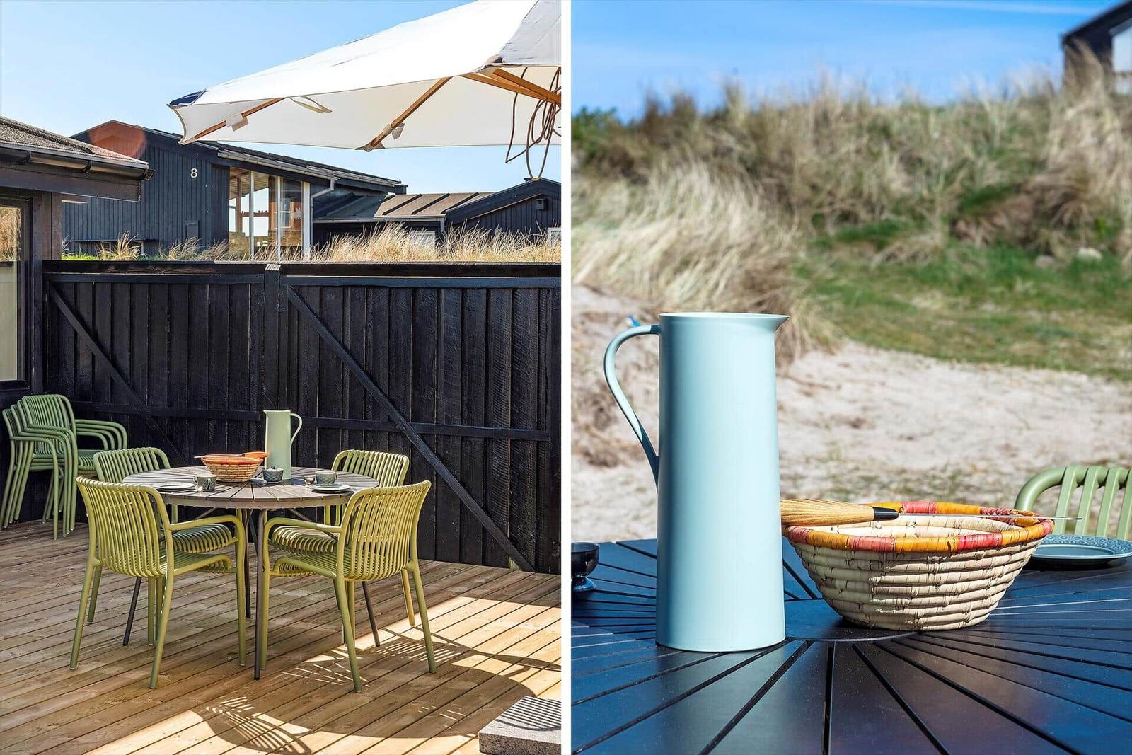 Terrasse mit Tisch, Stühlen und Sonnenschirm. Hintergrund: Strand und Dünen.