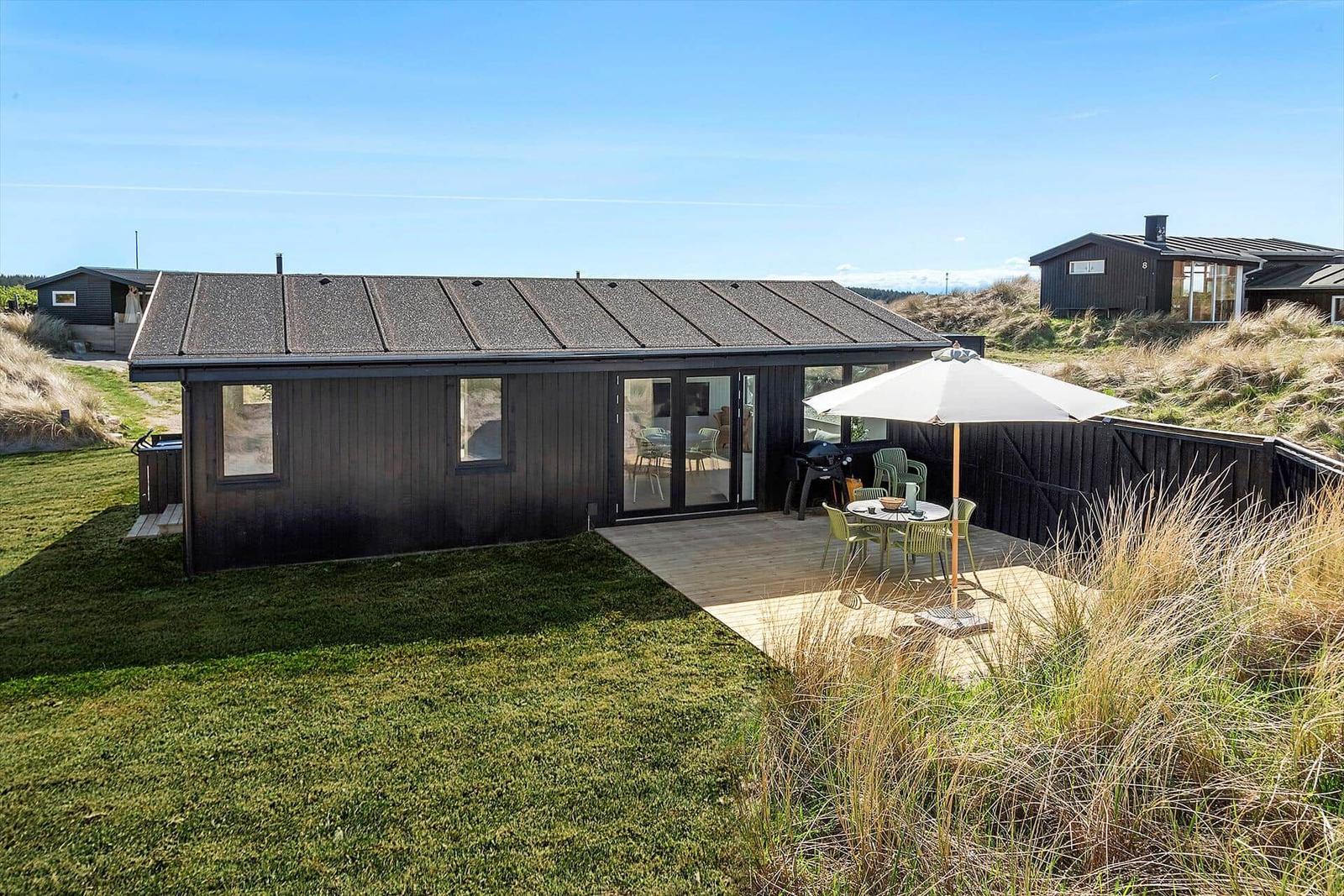 Moderne Hütte mit Terrasse, Garten und Grillplatz. Blick auf Dünen und klaren Himmel.