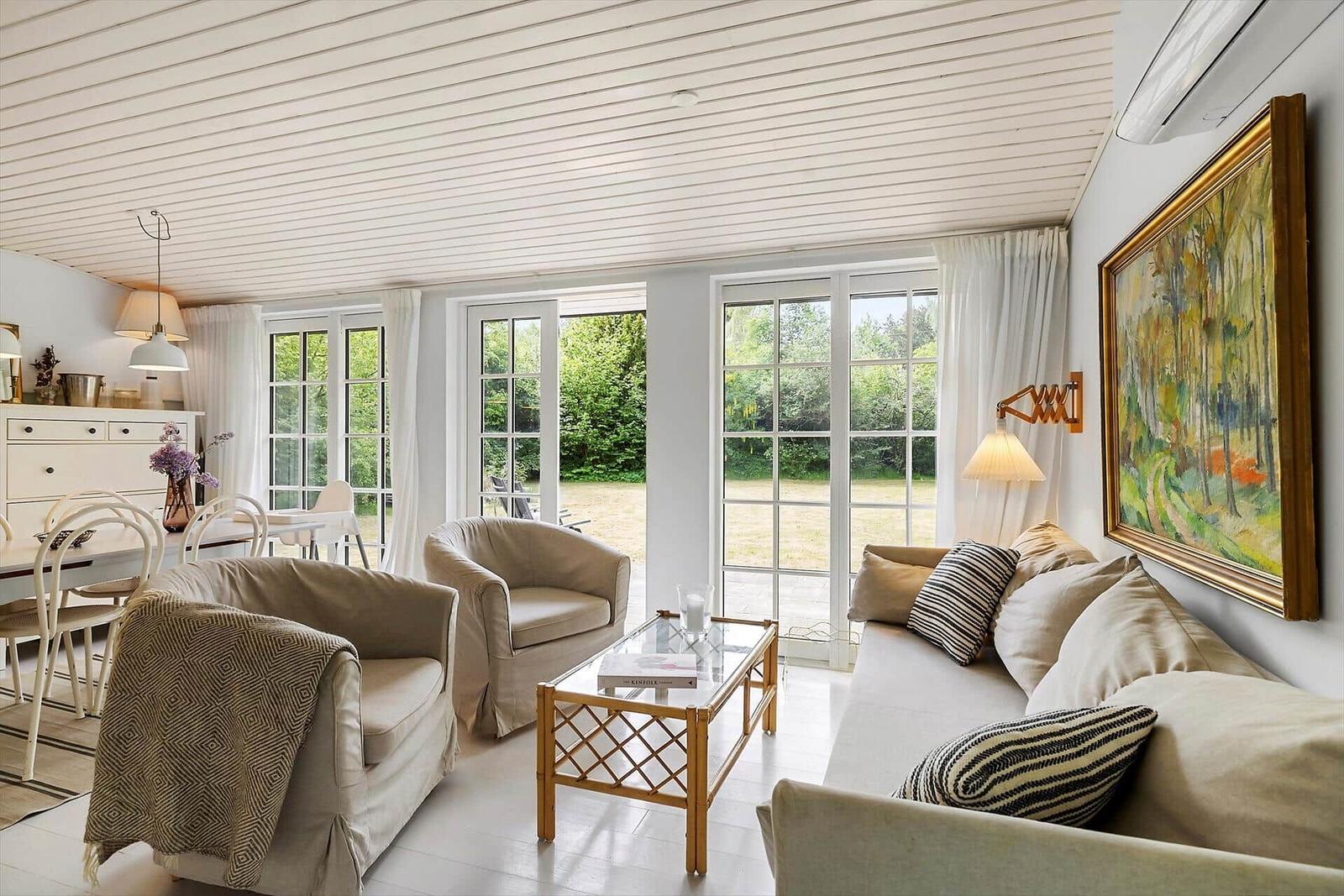 Living and dining area with sofa, armchairs, and glass table. Doorways to garden. Ceiling with wood paneling.