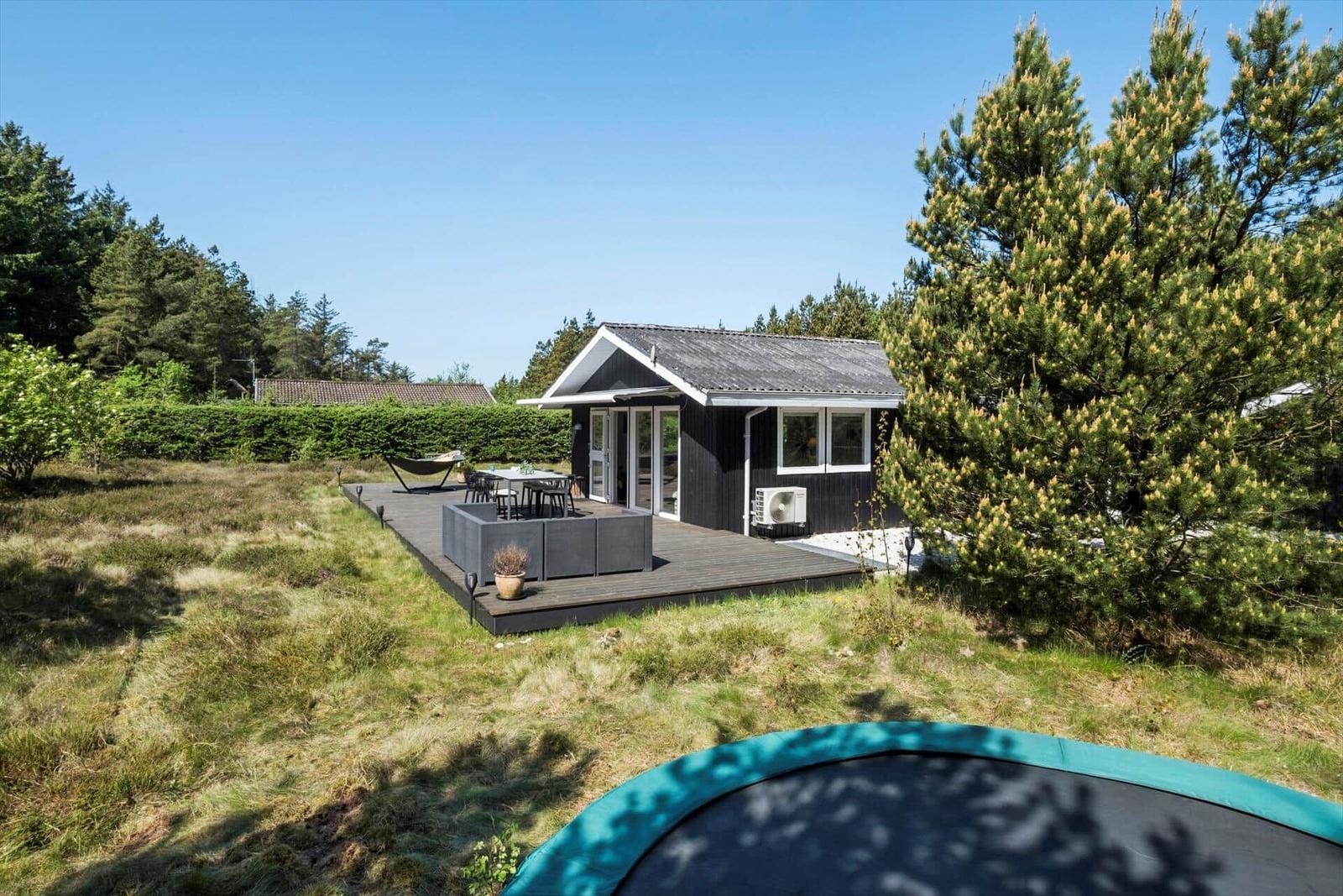 Schwarze Hütte mit Terrasse, Garten und Trampolin. Umgeben von Bäumen und Gras.