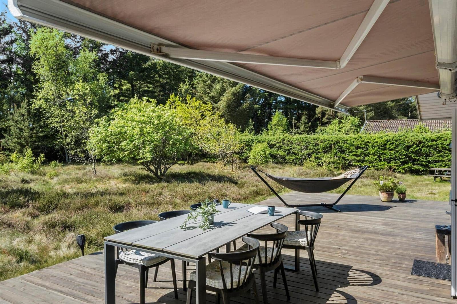 Terrasse mit Tisch, Stühlen und Hängemöbel unter Markise. Grüner Garten im Hintergrund.