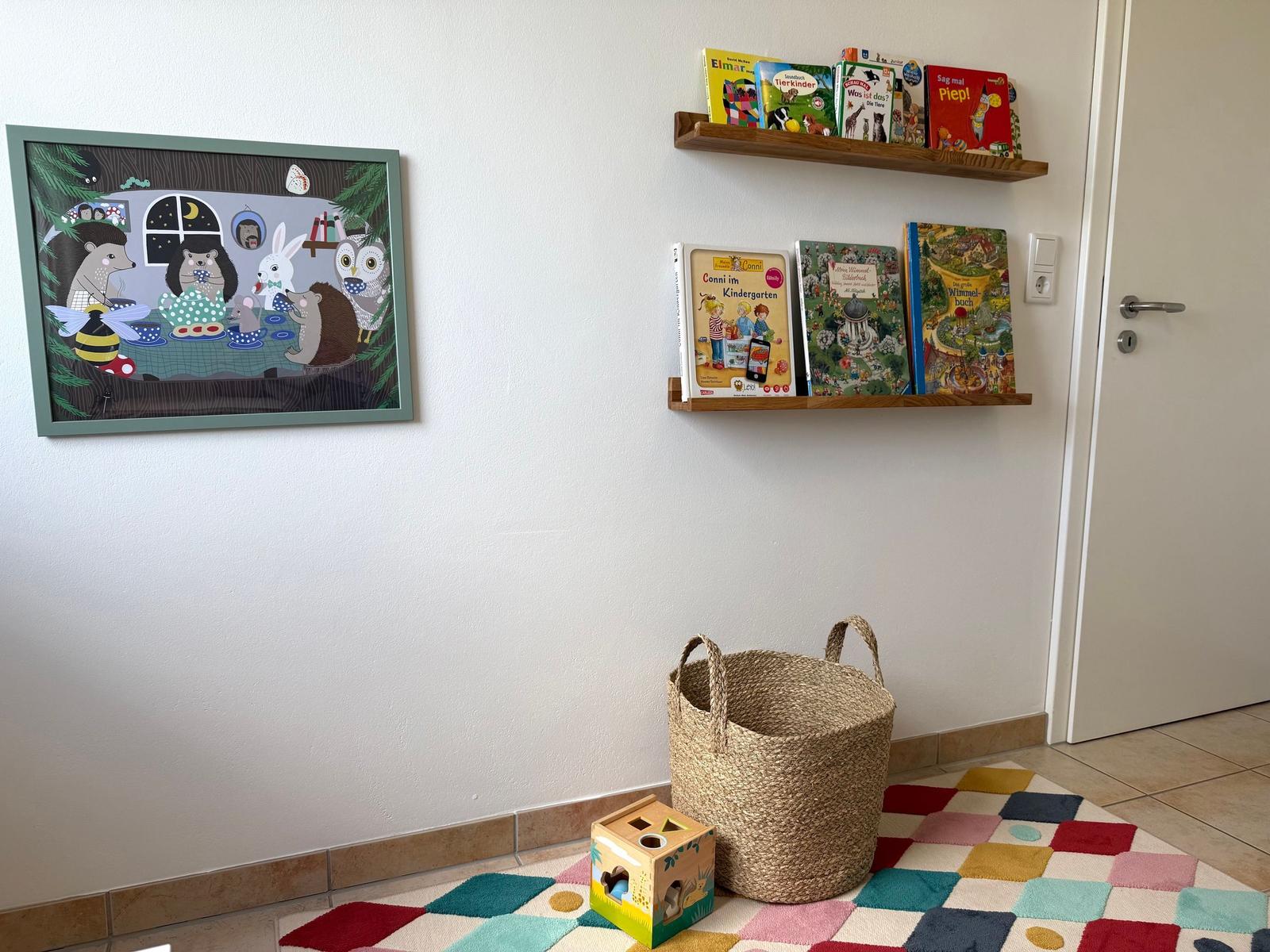 Wand mit Bildern, Bücherregal und Kinderspielzeug auf buntem Teppich.