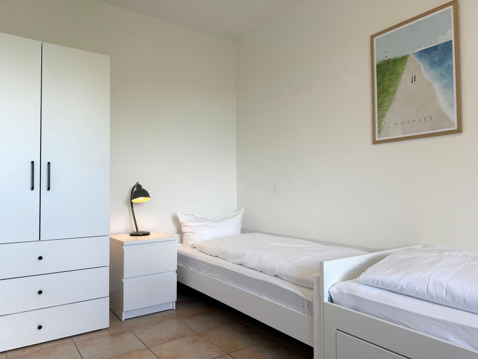 Zimmer mit zwei Betten, Schrank und Nachttisch mit Lampe.