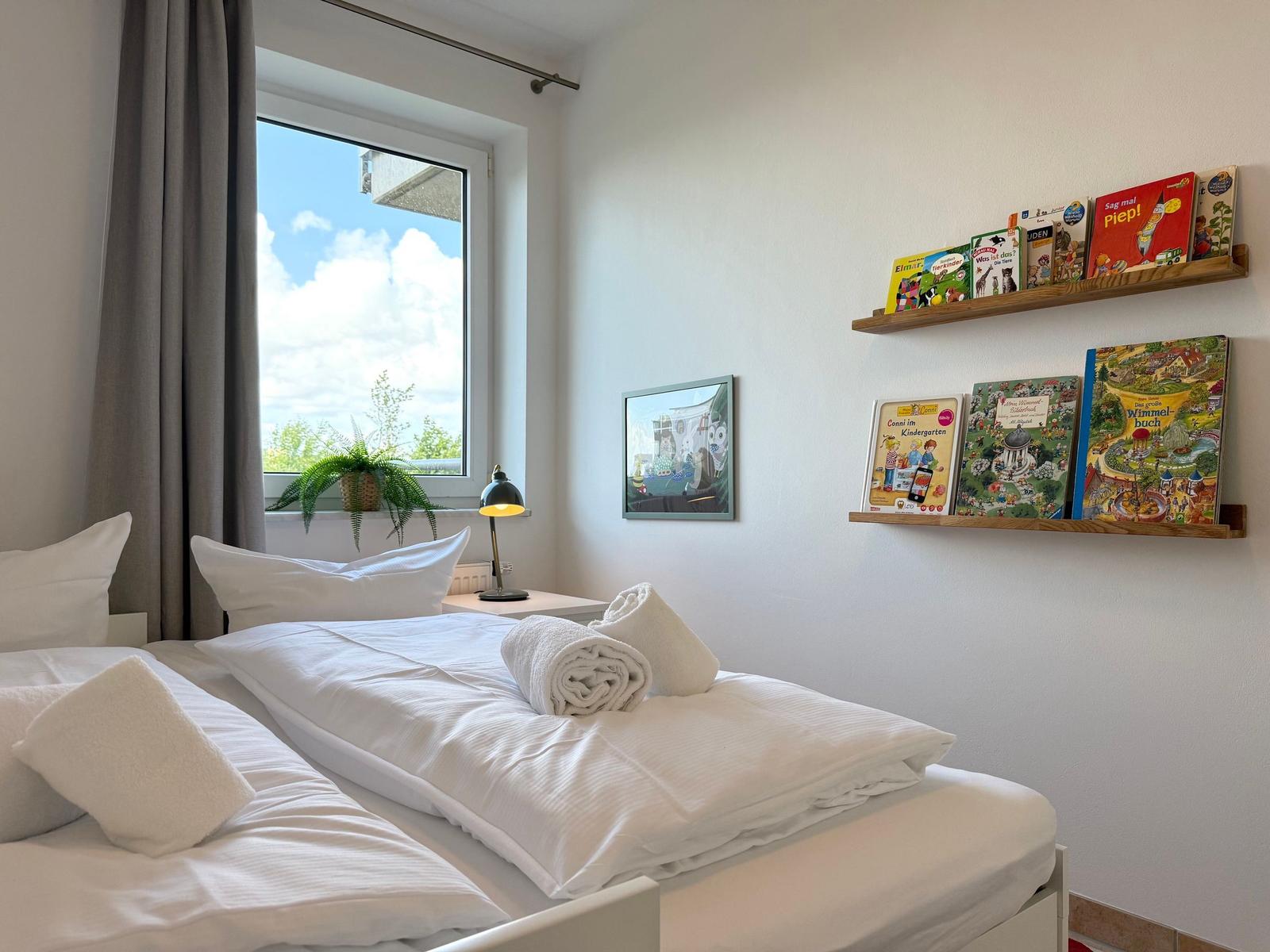 Schlafzimmer mit Bett, Fenster und Kinderbüchern auf Regalen.