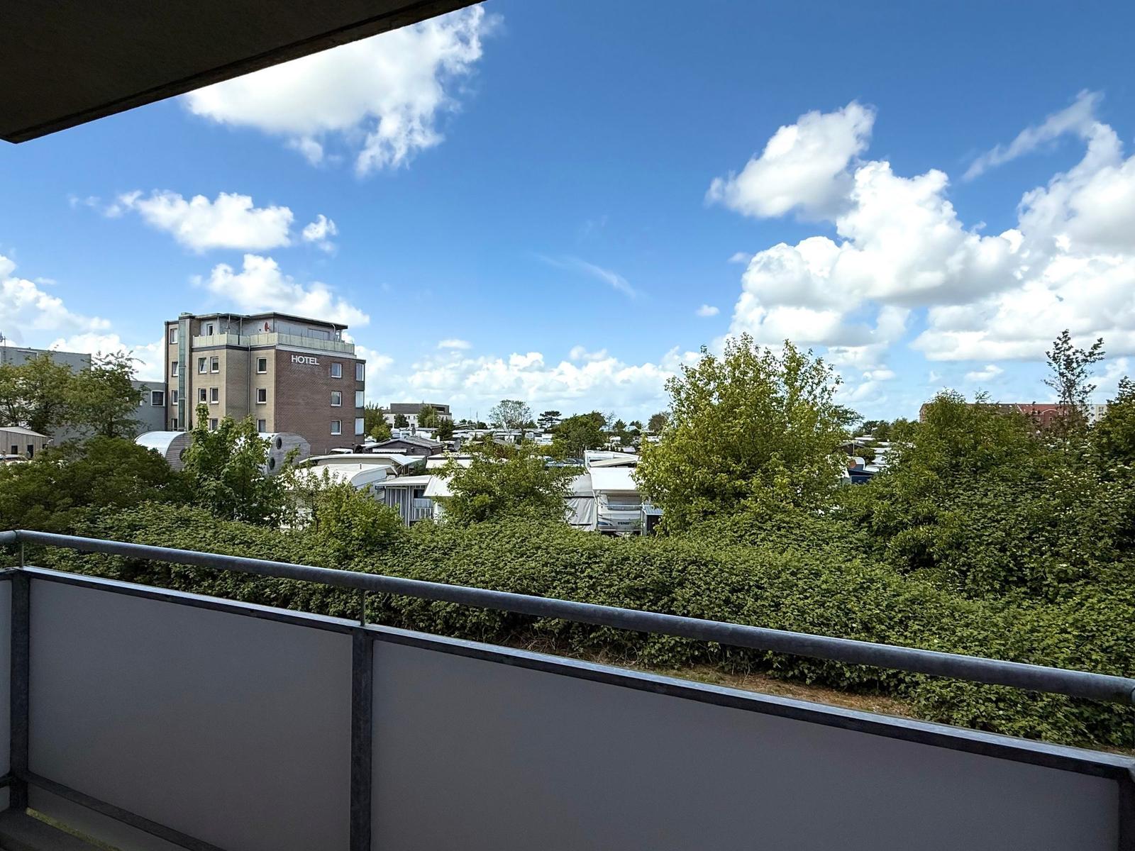 Balkon mit Blick auf Hotel und grüne Bäume unter blauem Himmel mit weißen Wolken.