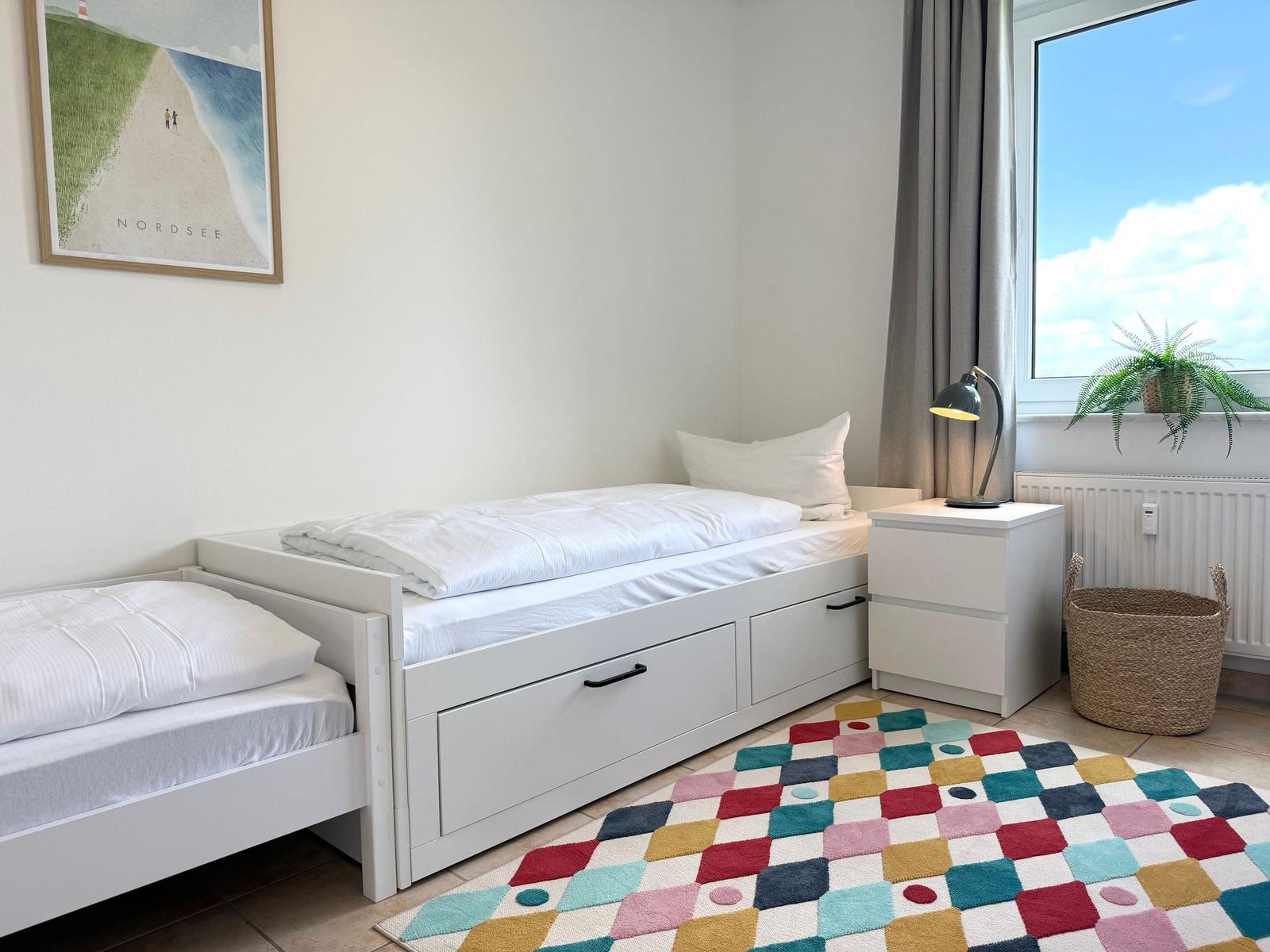 Zimmer mit Doppelbett, Nachttisch und buntem Teppich. Fenster mit Pflanze und Vorhang.