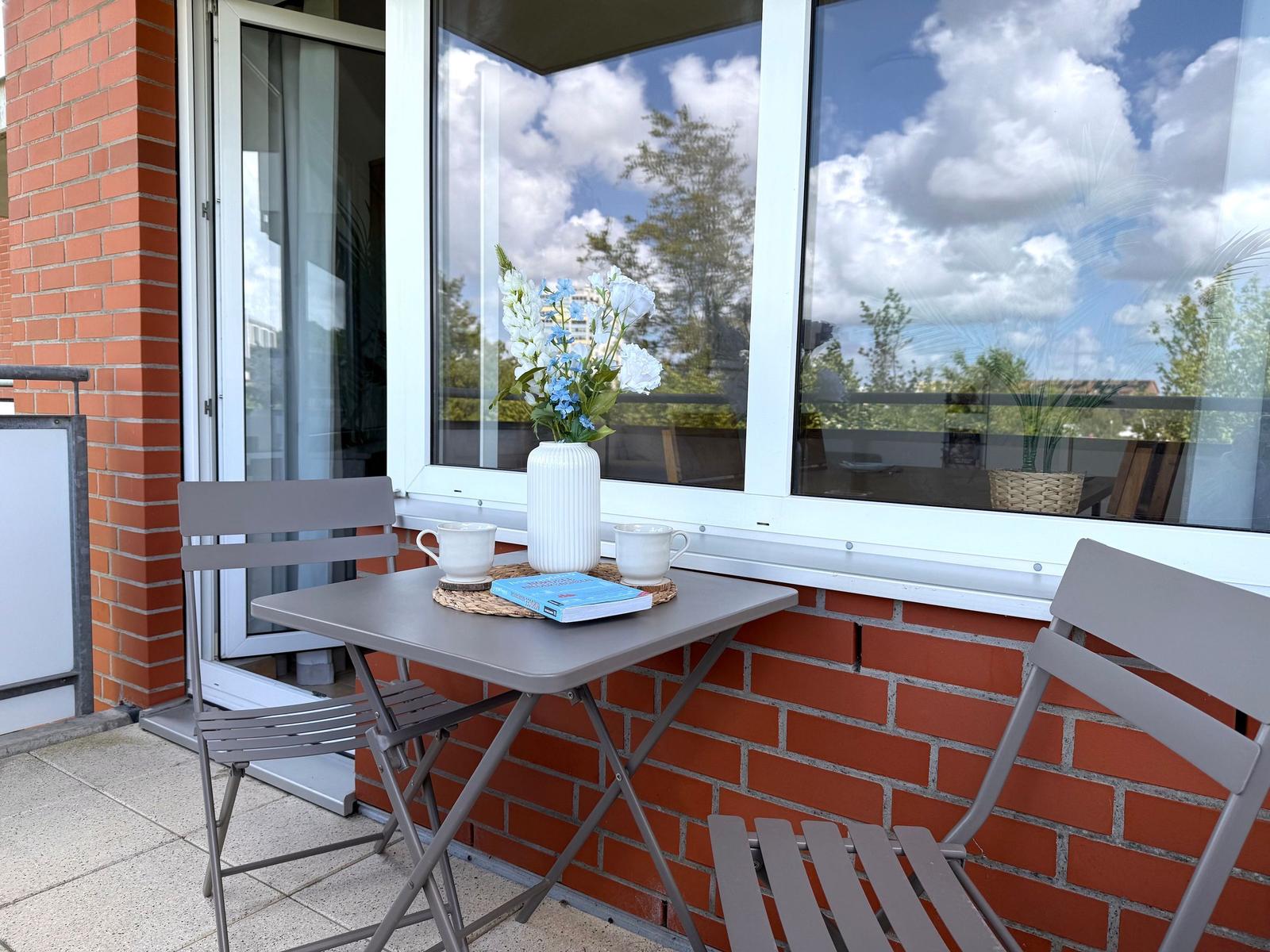 Balkon mit Tisch, zwei Stühlen und Blumenvase. Hintergrund: Glasfront und rote Ziegelwand.