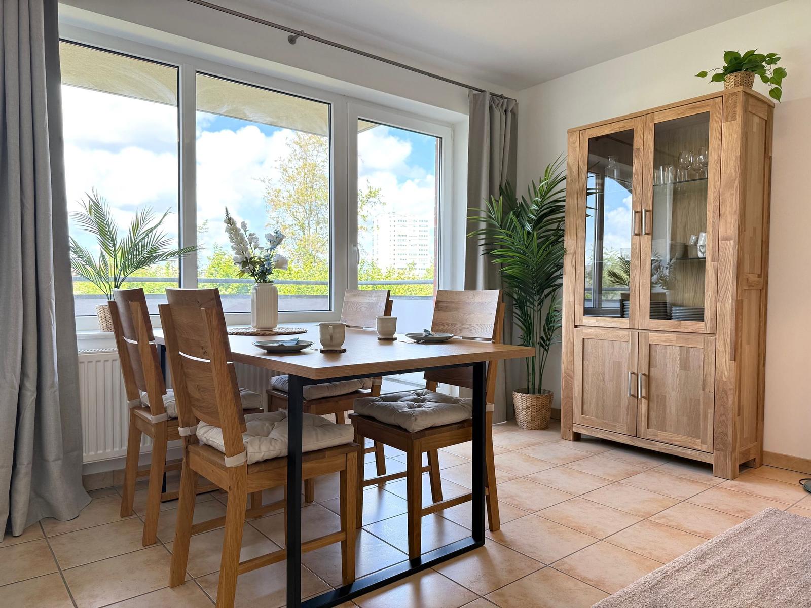 Essbereich mit Holztisch, Stühlen und Glaswand-Schrank. Große Fenster mit Blick auf Balkon.
