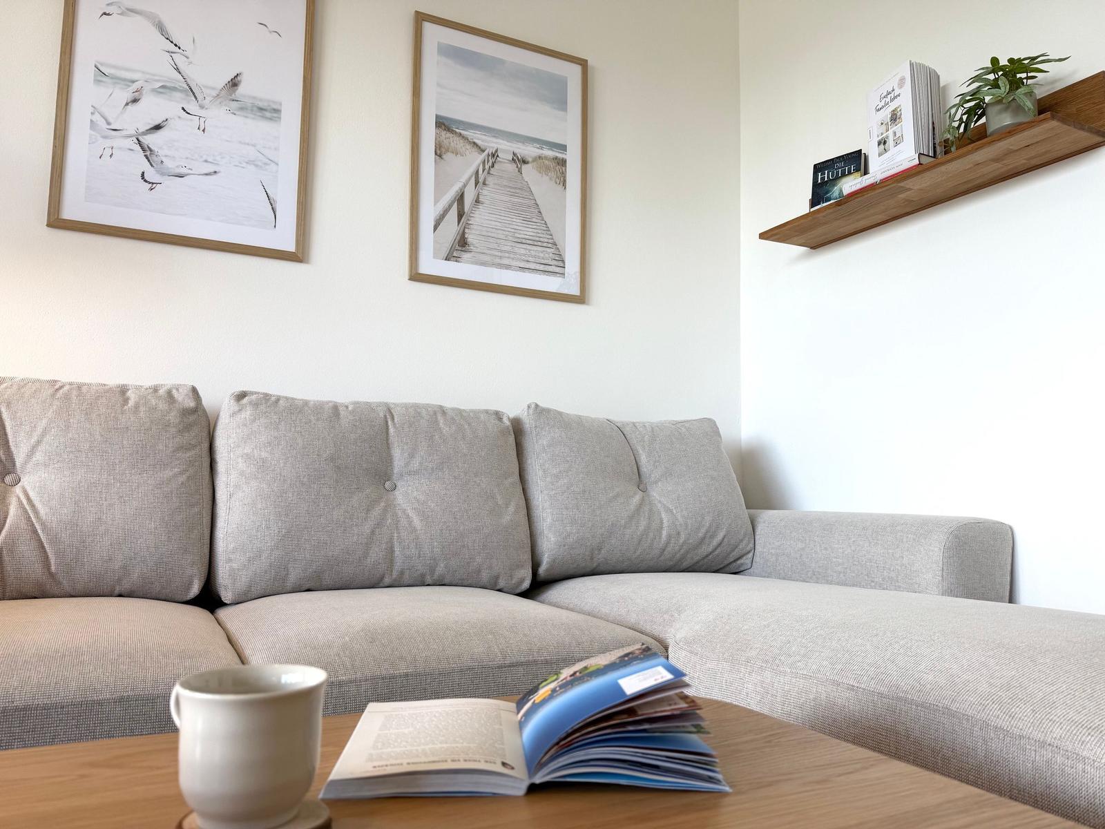 Ein grauer Sofa mit Kissen, Wandbilder und ein Tisch mit Buch und Tasse.