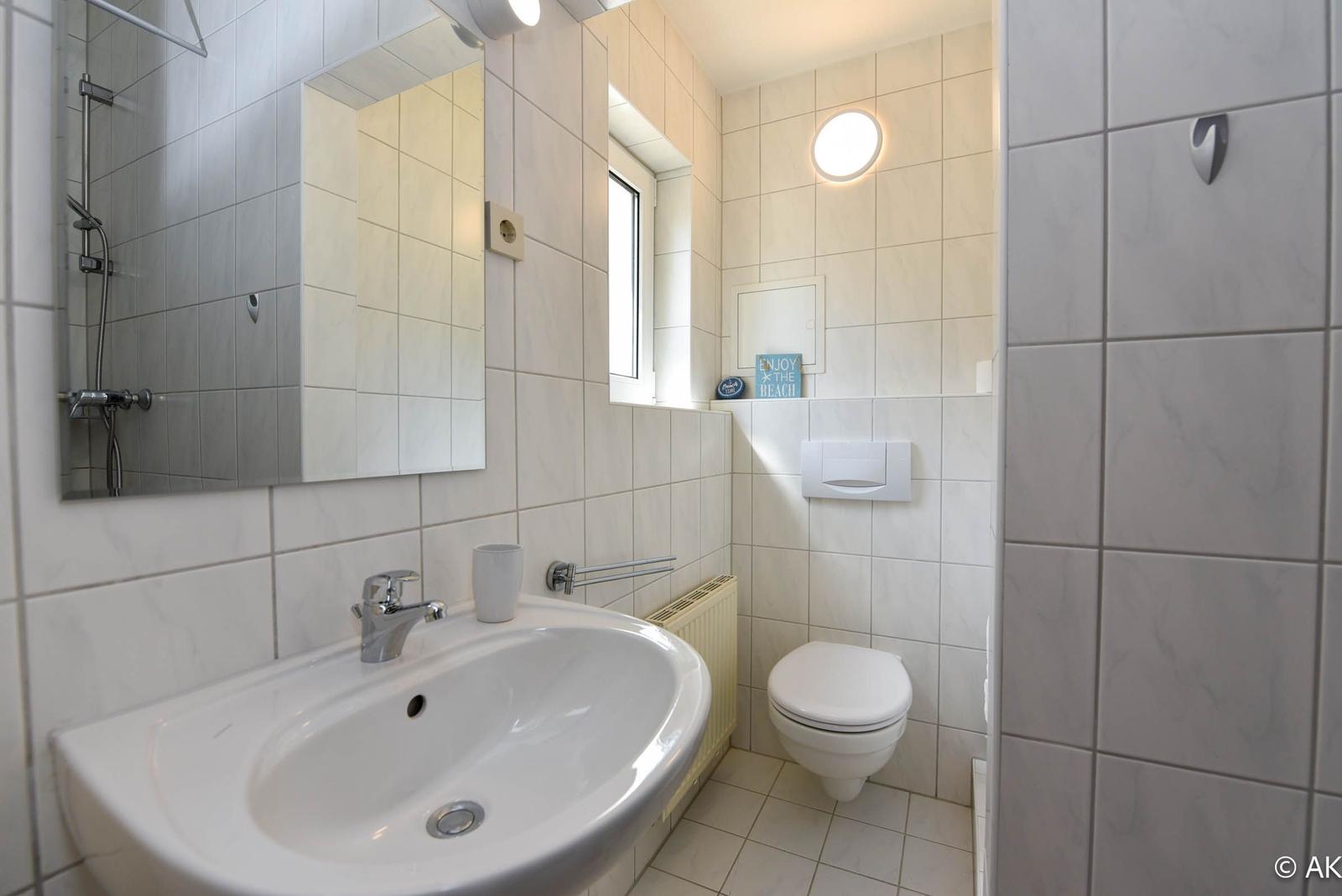 Badezimmer mit Dusche, Waschbecken und WC. Weiße Fliesen und Fenster.