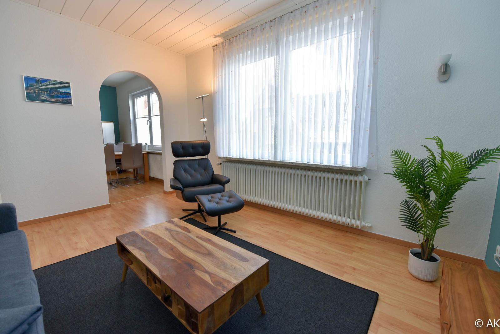 Wohnzimmer mit Sitzgelegenheit, Holztisch und Pflanze. Durchgang zur Küche.