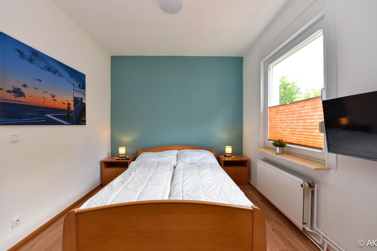 Schlafzimmer mit Doppelbett, Nachttischen und Fenster. Wandbild und Fernseher an der Wand.
