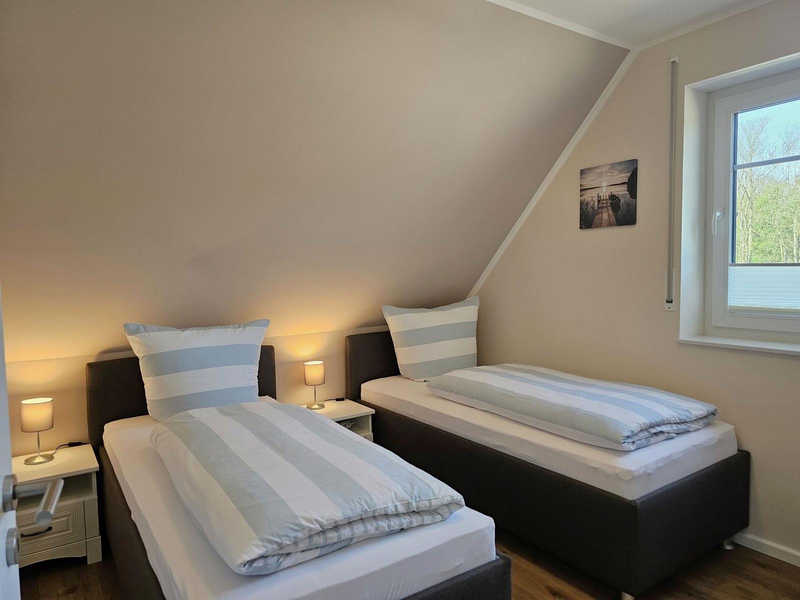 Zwei Einzelbetten mit Streifenbetten, Nachttische und ein Fenster mit Blick auf Bäume.