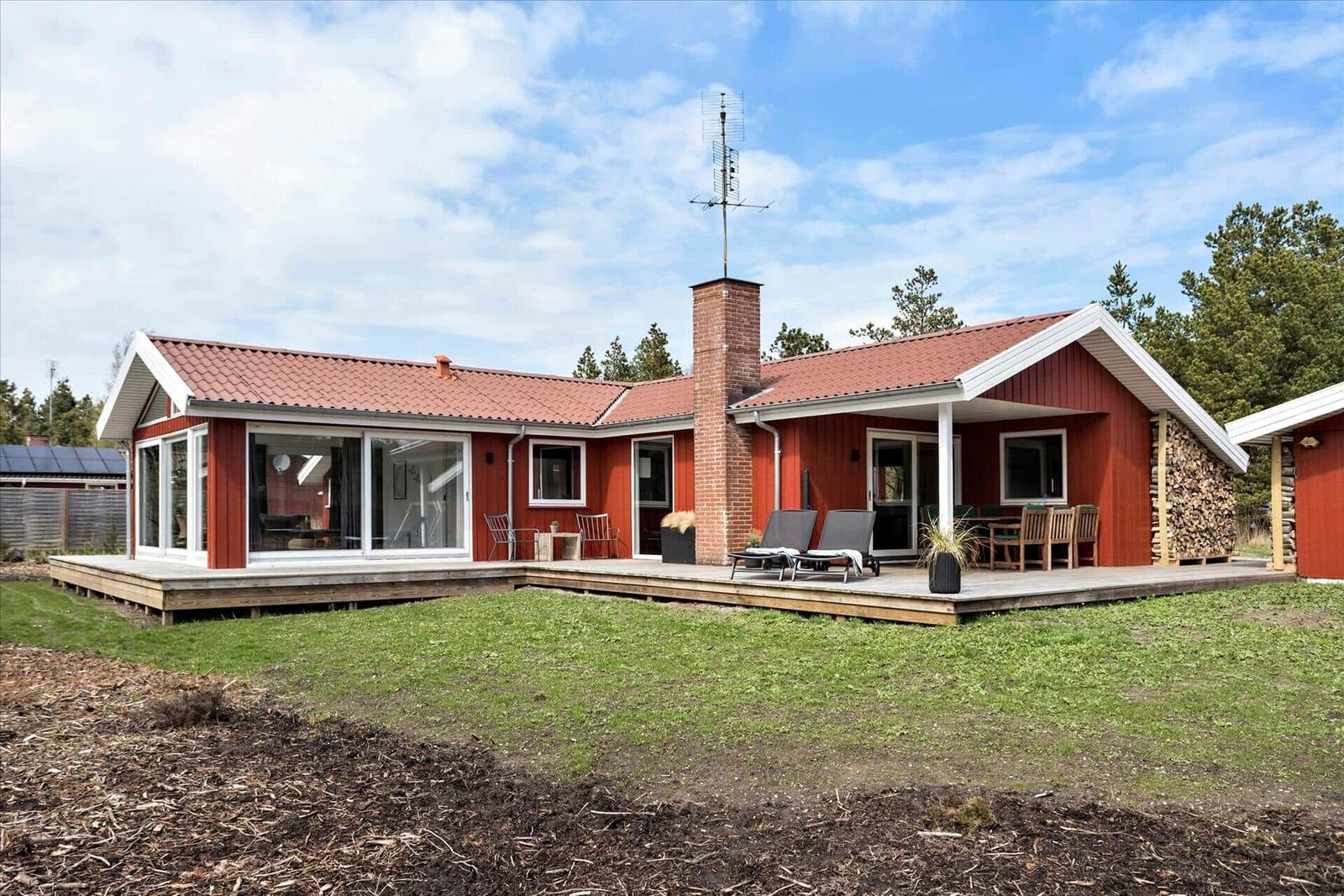Rotes Haus mit Terrasse, Liegen und Holzstapel. Grünfläche und Bäume umgeben.