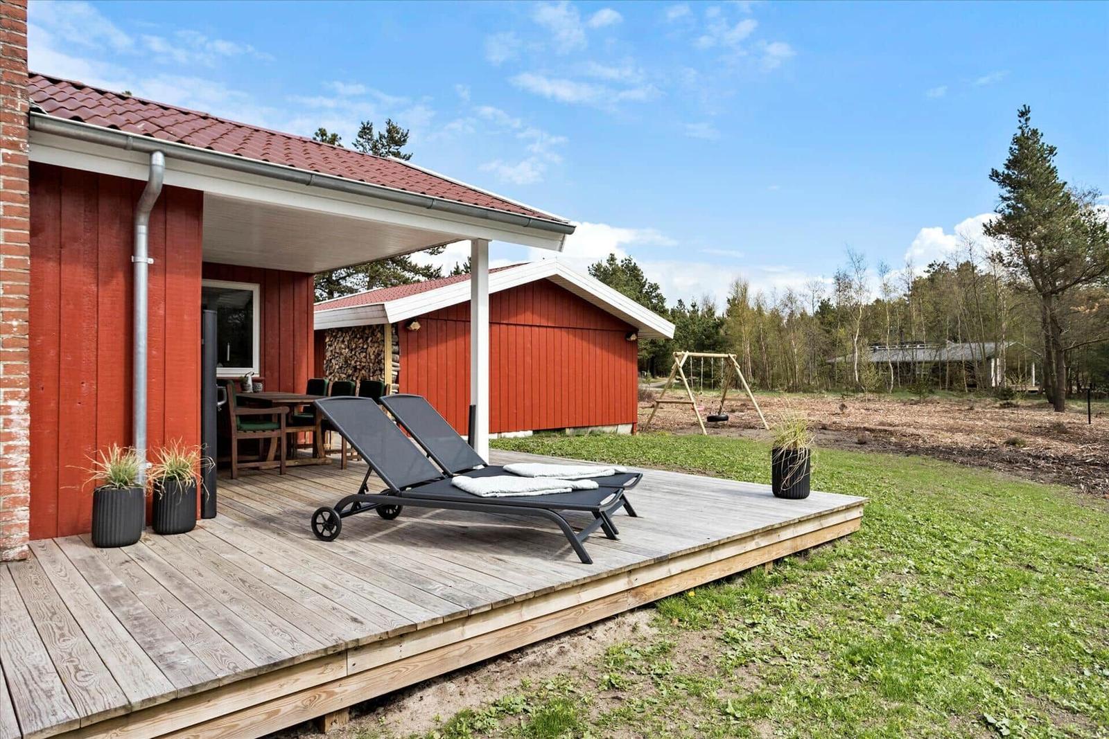 Terrasse mit Liegestühlen und Tisch unter Dach. Hinterhof mit Spielgeräten und Wald.