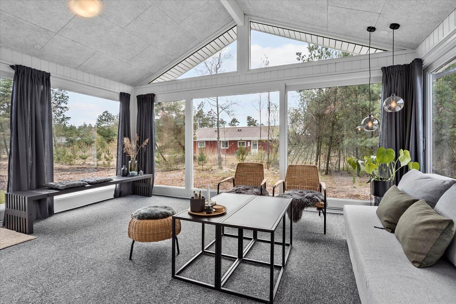 Ein Wohnraum mit großem Fenster, Sofa, Tisch und Sitzgelegenheiten. Blick auf Wald und Haus.