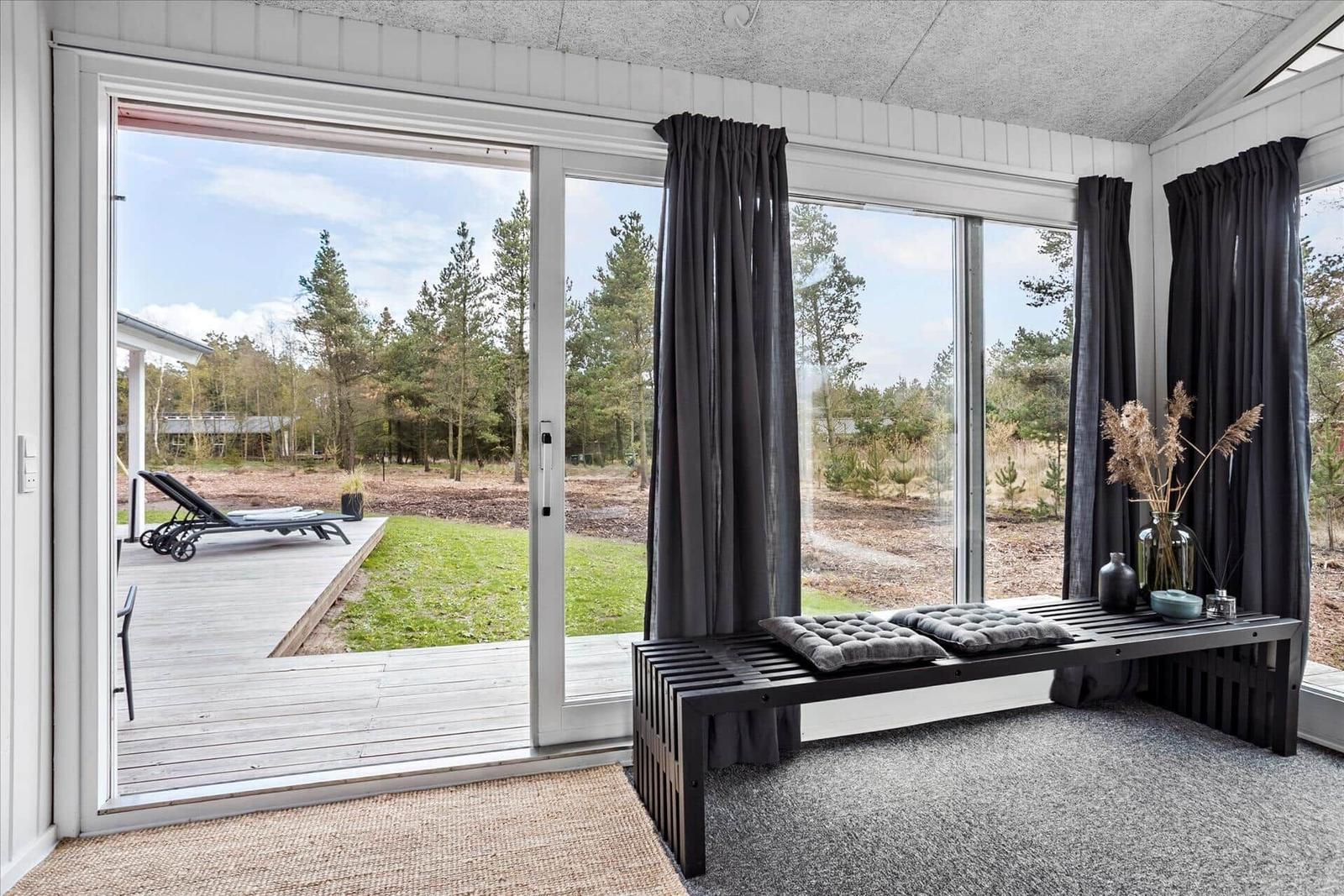 Raum mit großem Fenster, Blick auf Terrasse und Wald. Schwarze Gardinen und Bank mit Kissen.