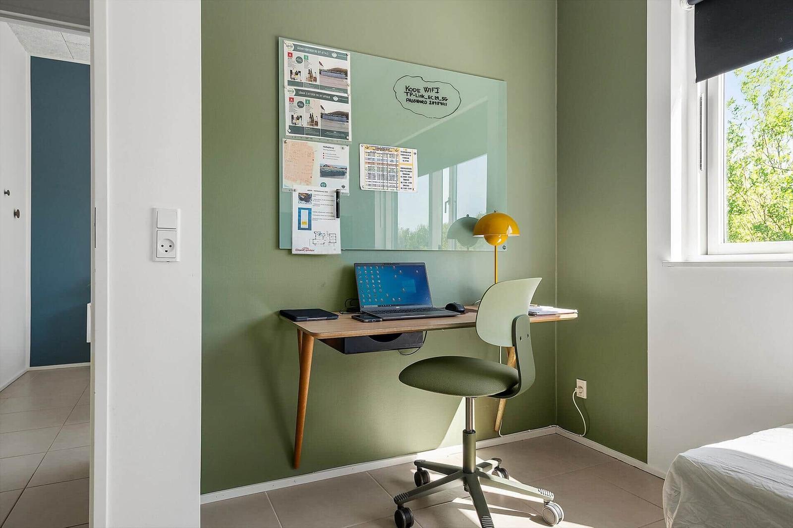 Arbeitsplatz mit Schreibtisch, Stuhl und Laptop an grüner Wand mit Fenster.