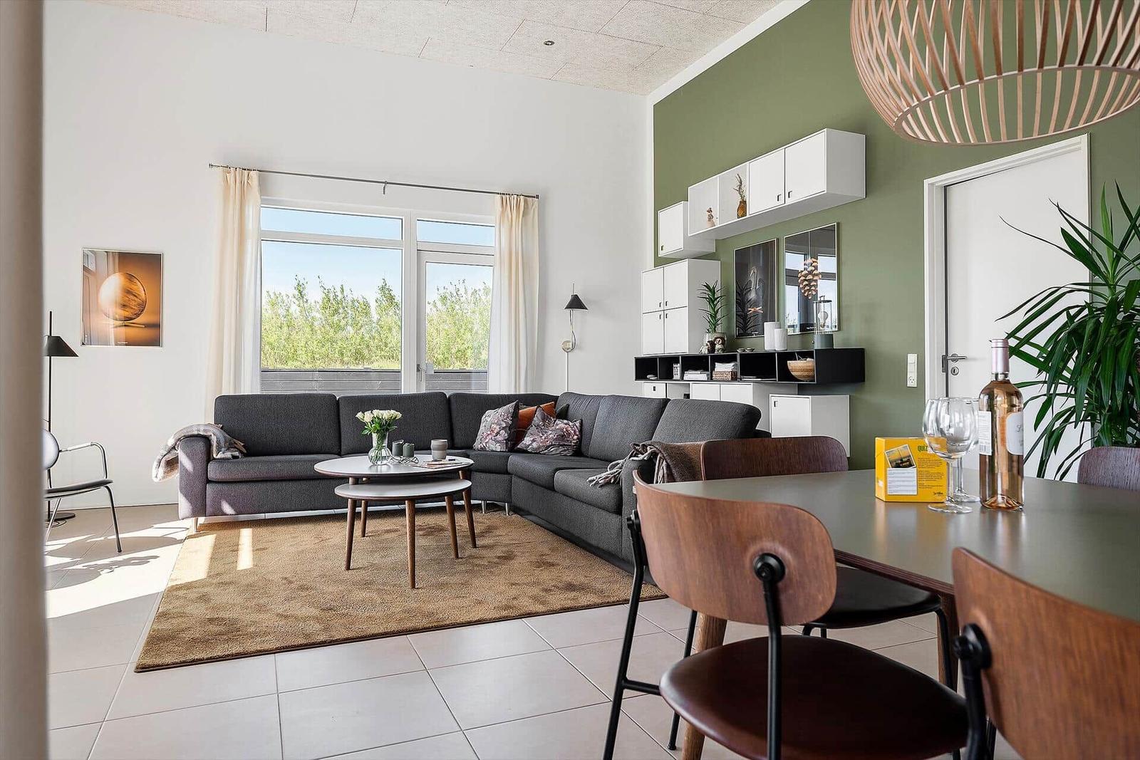 Wohn- und Essbereich mit Sofa, Tisch und Stühlen. Grüne Wand, Fenster und Pflanzen.