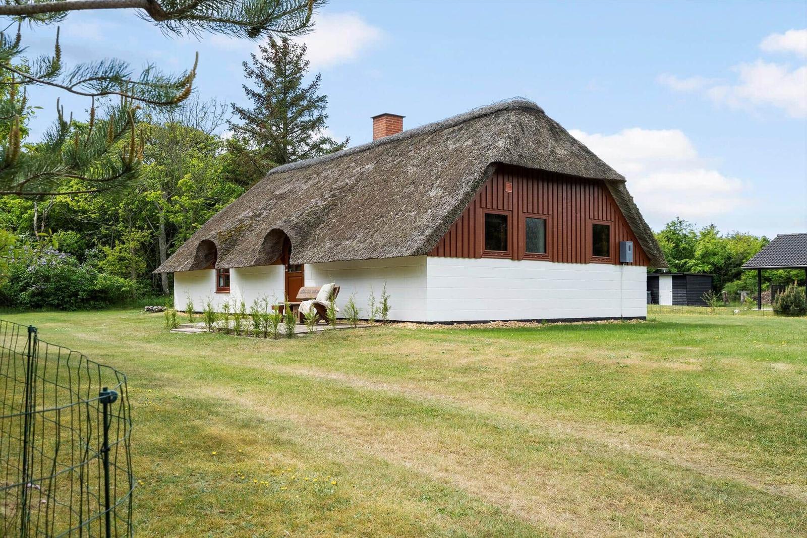 Haus mit Strohdach, weißer Fassade und Holzverkleidung, umgeben von Grünflächen und Bäumen.