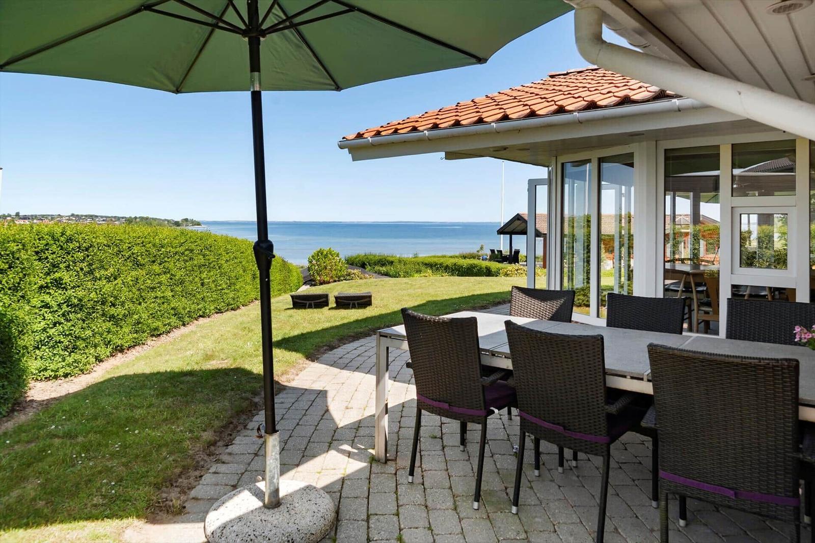 Terrasse mit Tisch, Stühlen und Sonnenschirm. Blick auf Meer und grüne Hecke.