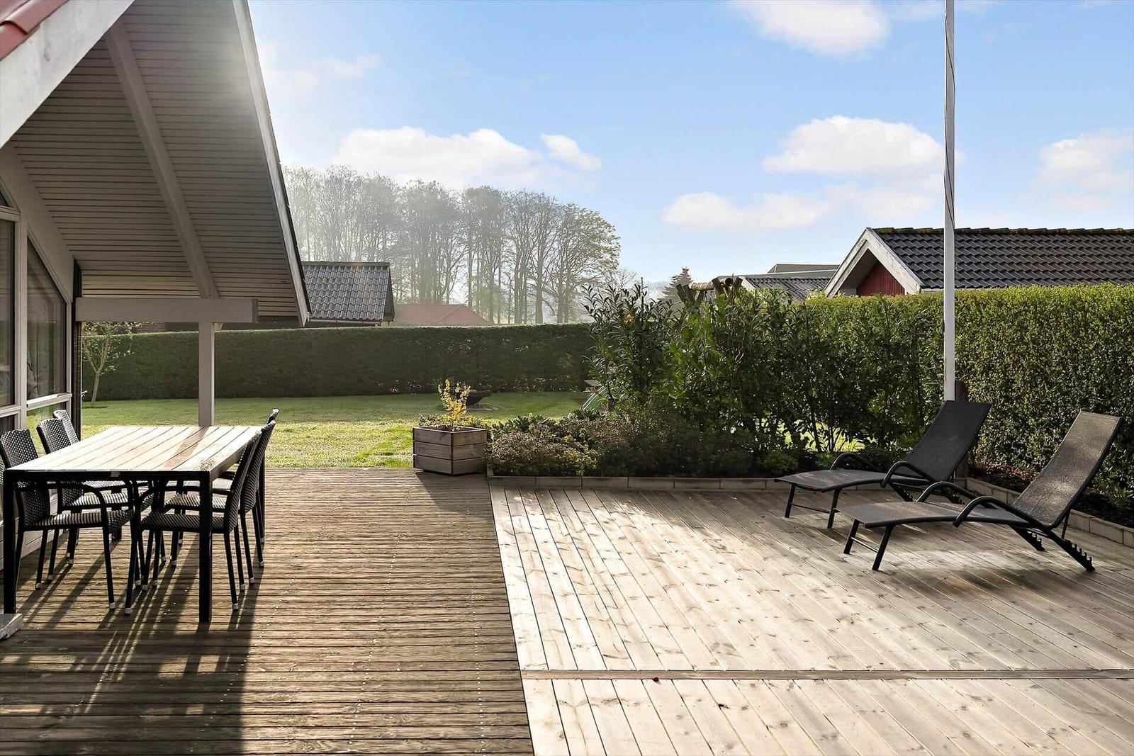 Wooden terrace with table, chairs, and lounge chairs. Lawn and hedge in the background.