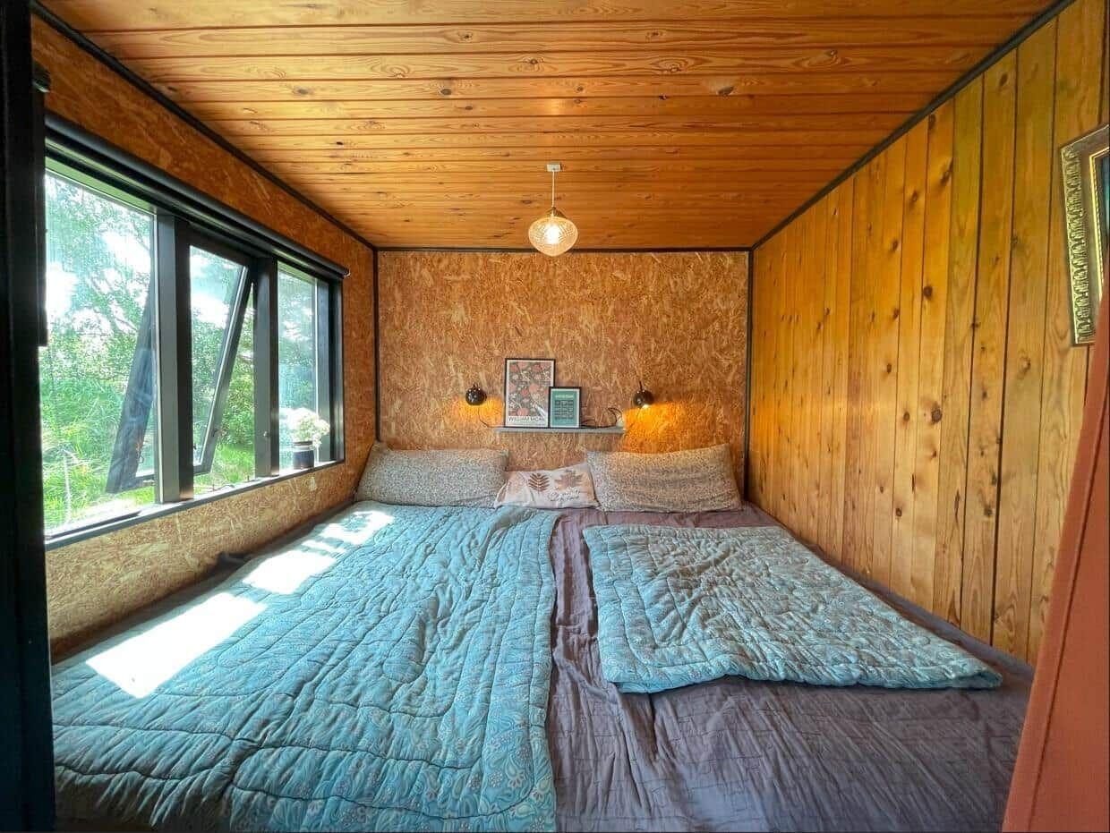 Schlafzimmer mit Doppelbett, Holzwand und großem Fenster zur Natur.