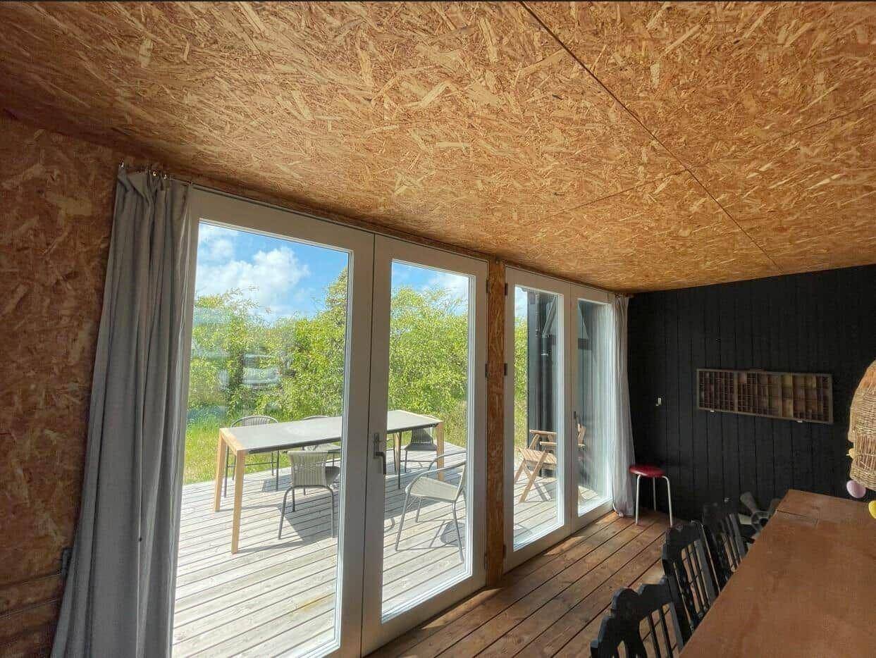 Raum mit Holzdecke und großen Fenstern zum Balkon mit Tisch und Stühlen.