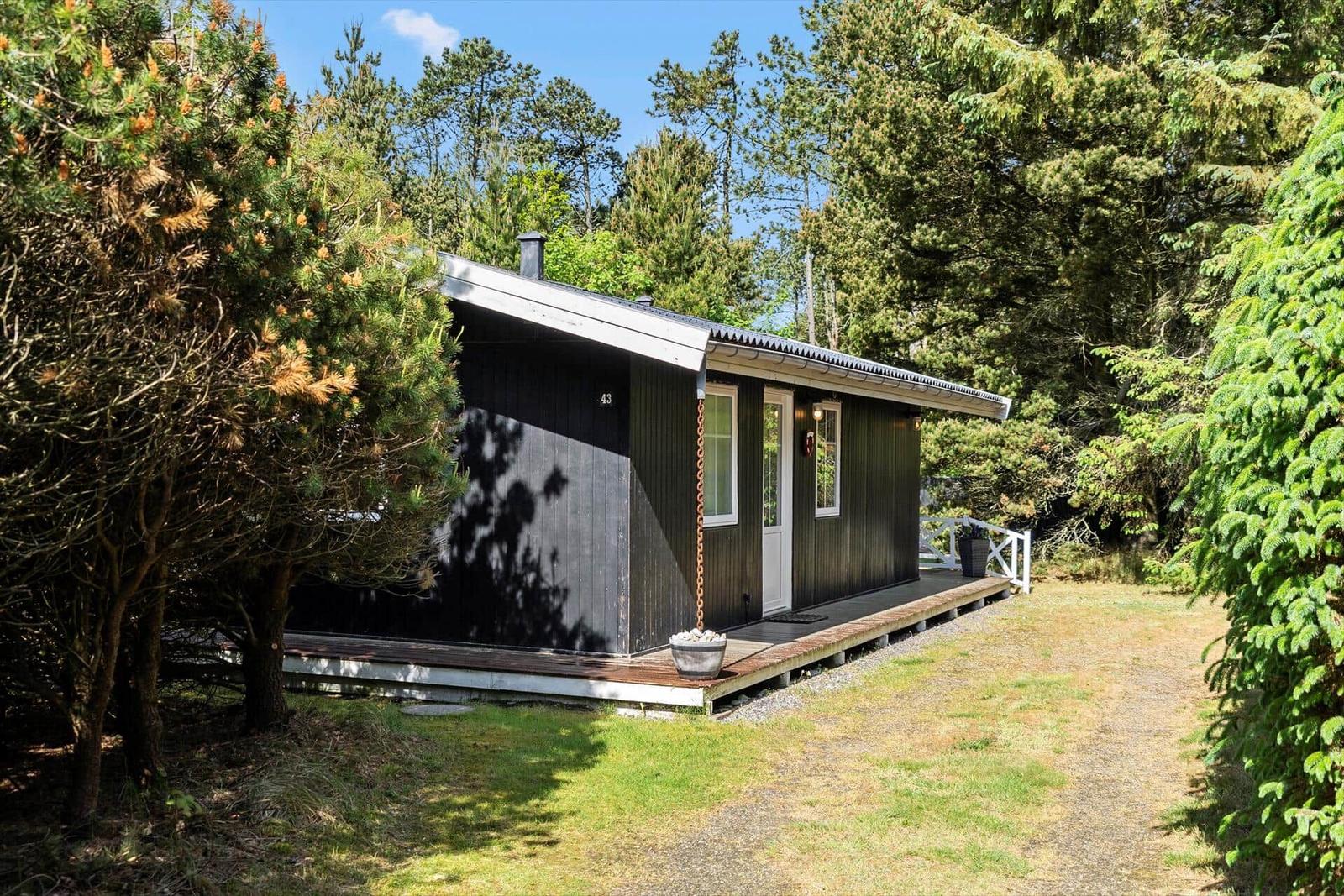 Ein kleines Haus mit dunkler Fassade und weißer Tür steht im Wald. Eine Terrasse führt zur Eingangstür.