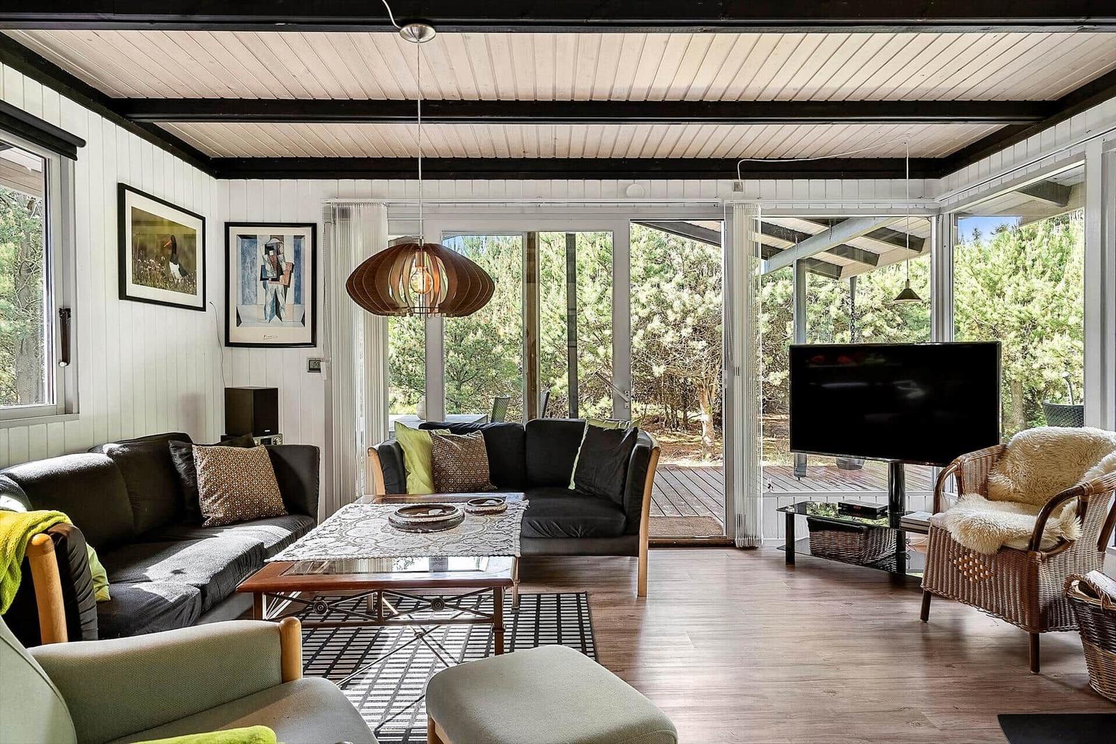 Wohnzimmer mit Sofa, Couchtisch und Fernseher. Große Fenster zeigen einen Garten.