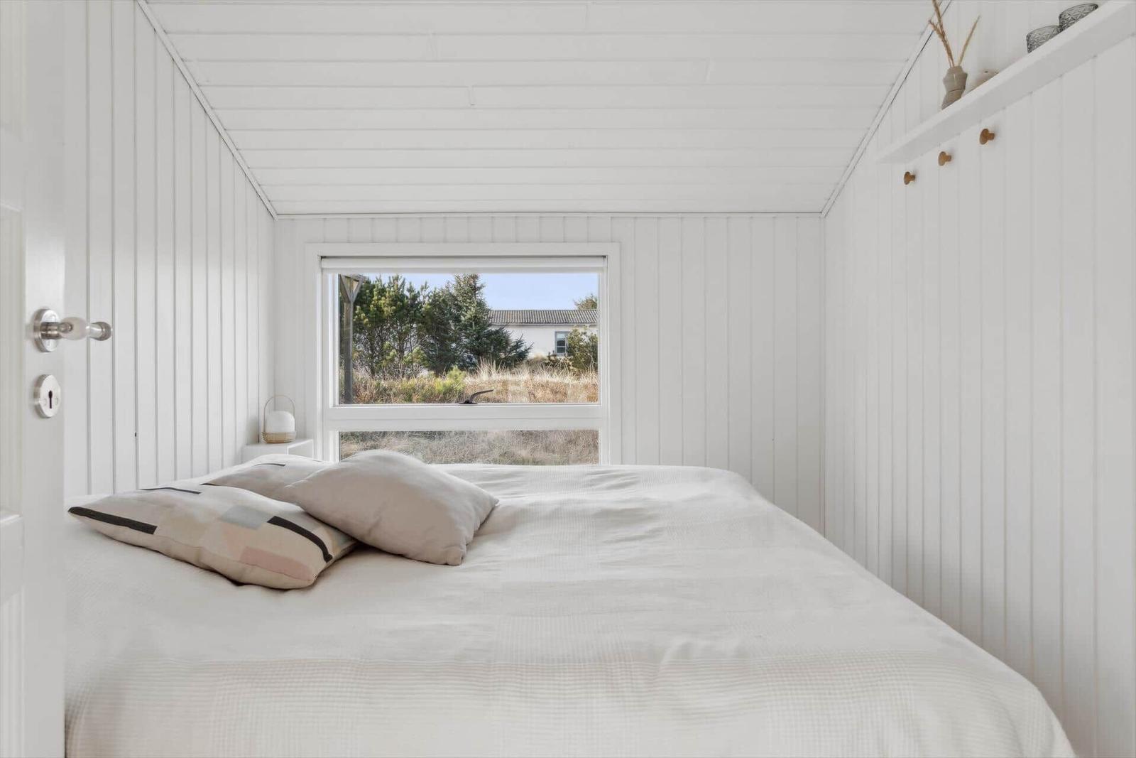 Bedroom with white wall paneling, bed, and view of green areas through window.