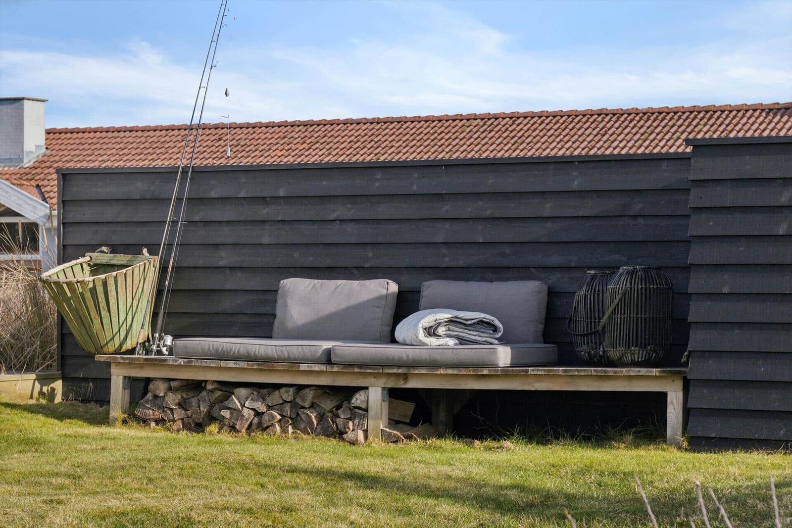 Outdoor bench with cushions and blanket in front of dark wooden house with chimney and tiled roof.