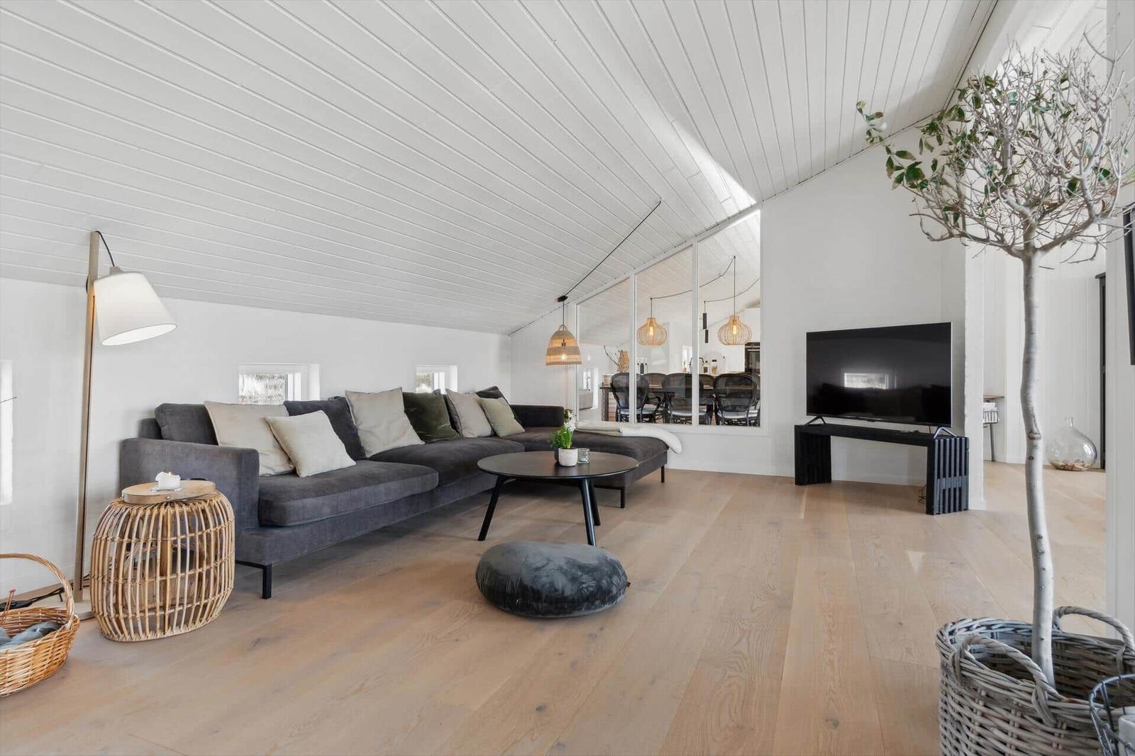 Living room with gray sofa, wooden floor, and white ceiling. TV and plant are visible.
