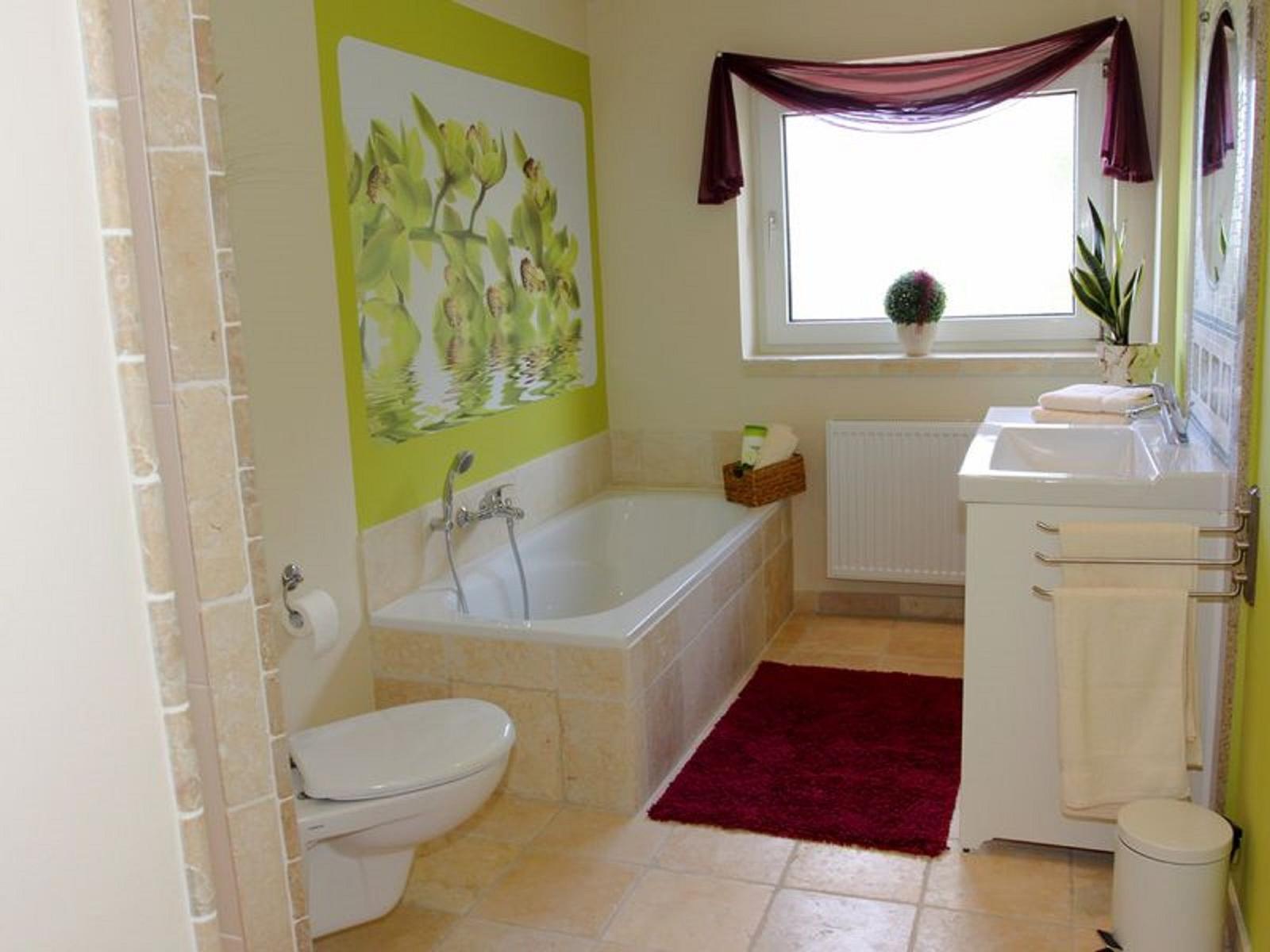 Badezimmer mit Badewanne, Waschbecken und Toilette. Wandbild und Fenster mit Vorhang.