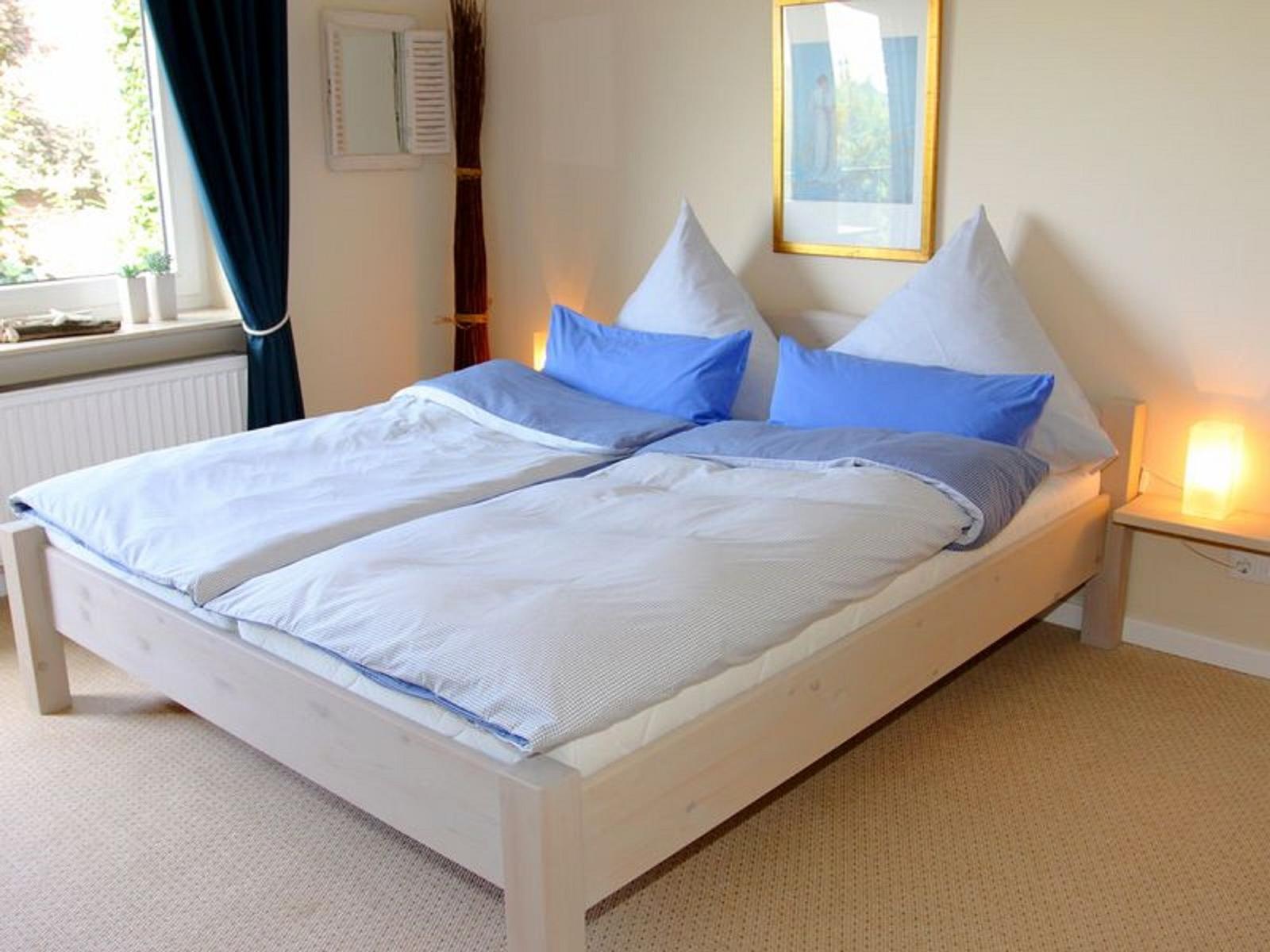 Doppelbett mit blauen Kissen und weißen Bettwäsche. Fenster mit dunklen Vorhängen und Nachttisch mit Lampe.