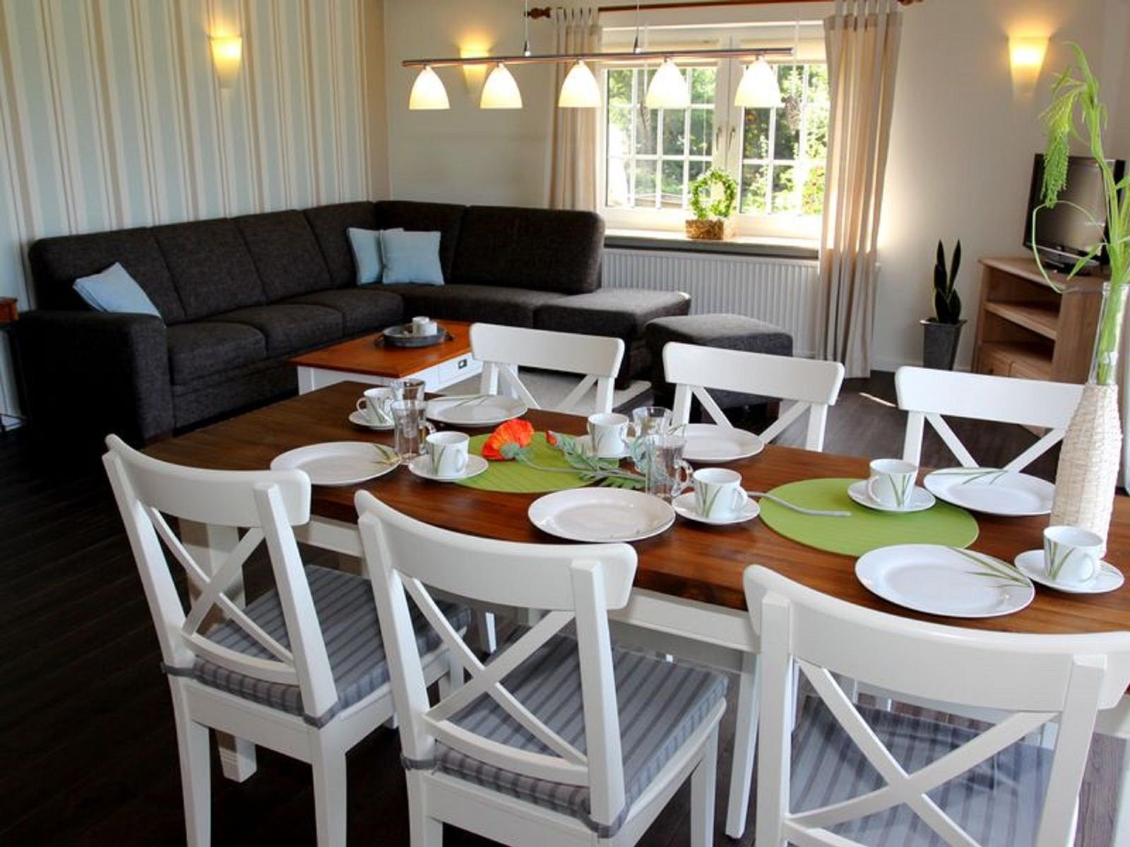 Esszimmer mit Holztisch, weißen Stühlen und dunklem Sofa im Hintergrund.