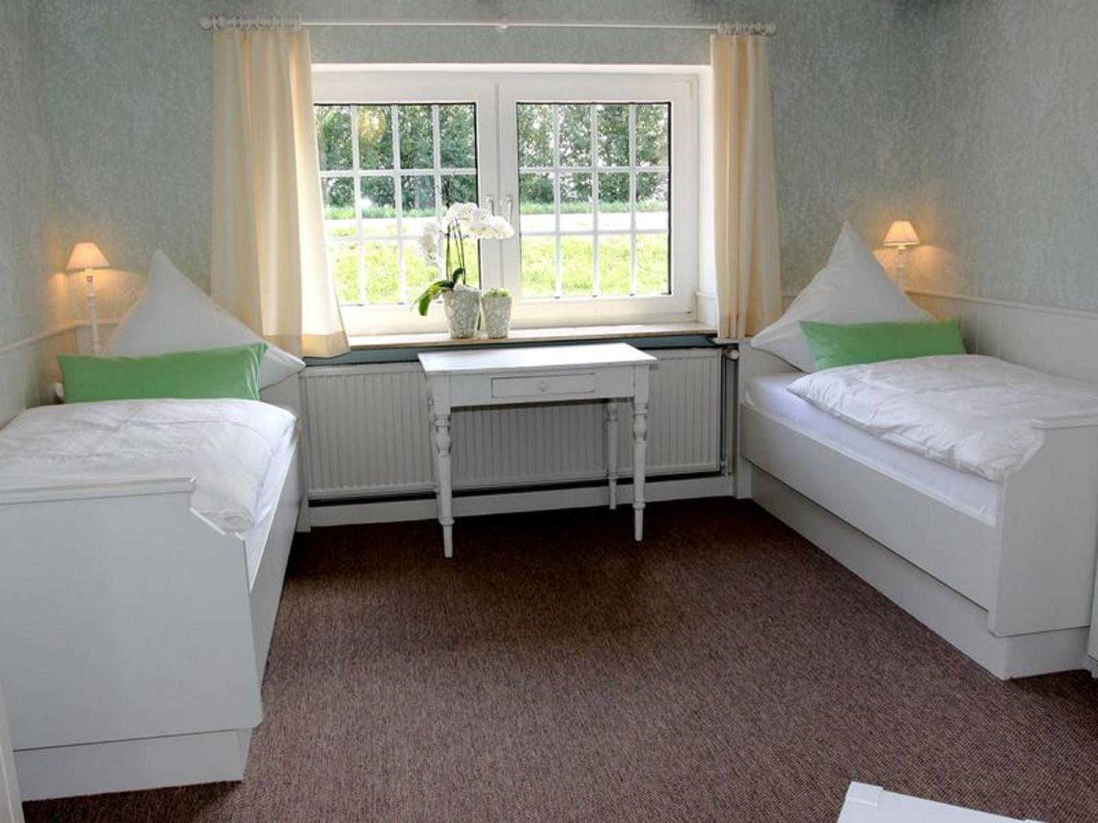Zwei Einzelbetten mit grünen Kissen, Fenster mit Vorhängen und Schreibtisch.