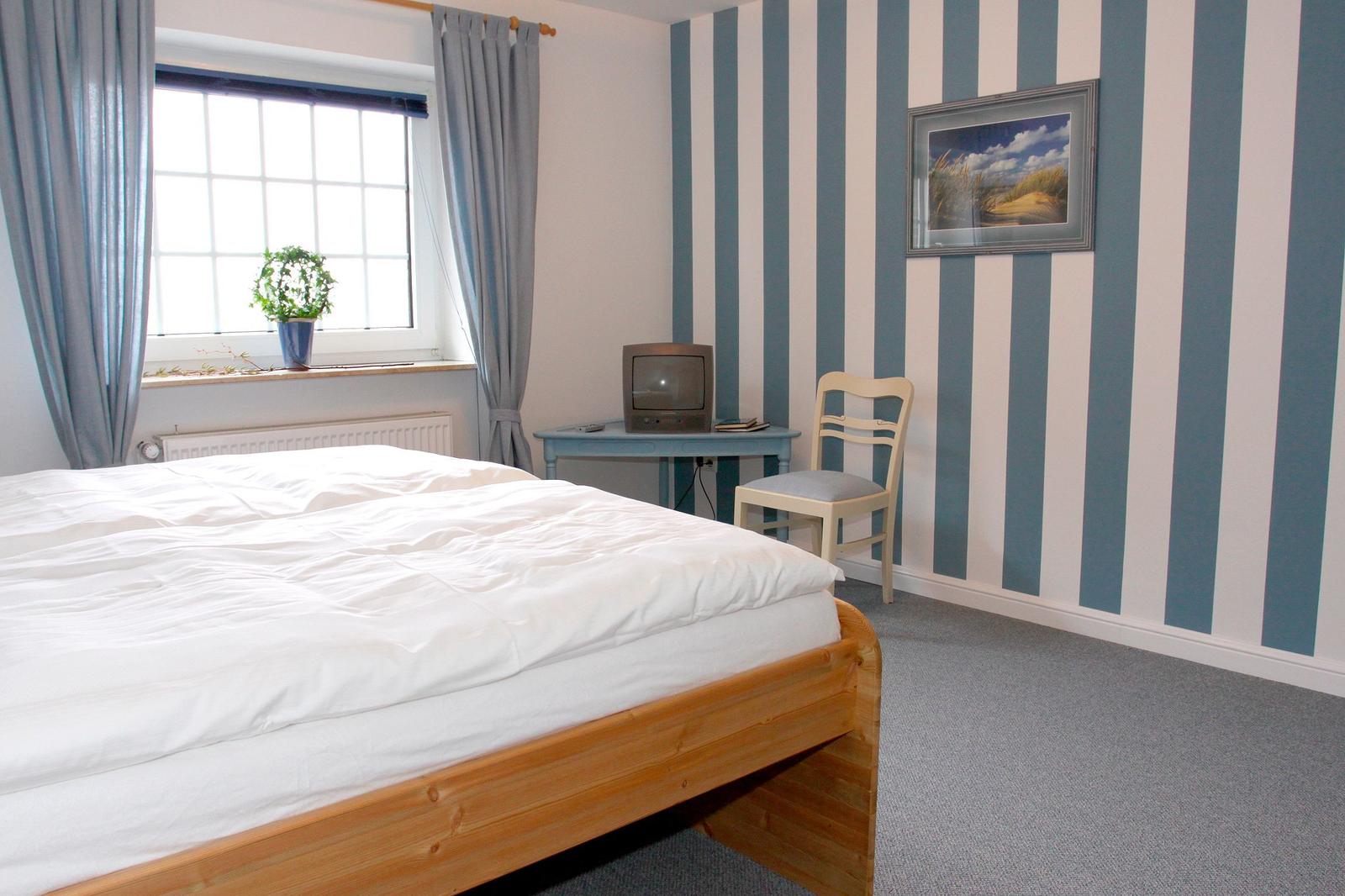 Ein Schlafzimmer mit Bett, Schreibtisch, Stuhl und Streifenwand. Fenster mit Vorhängen und Pflanze.