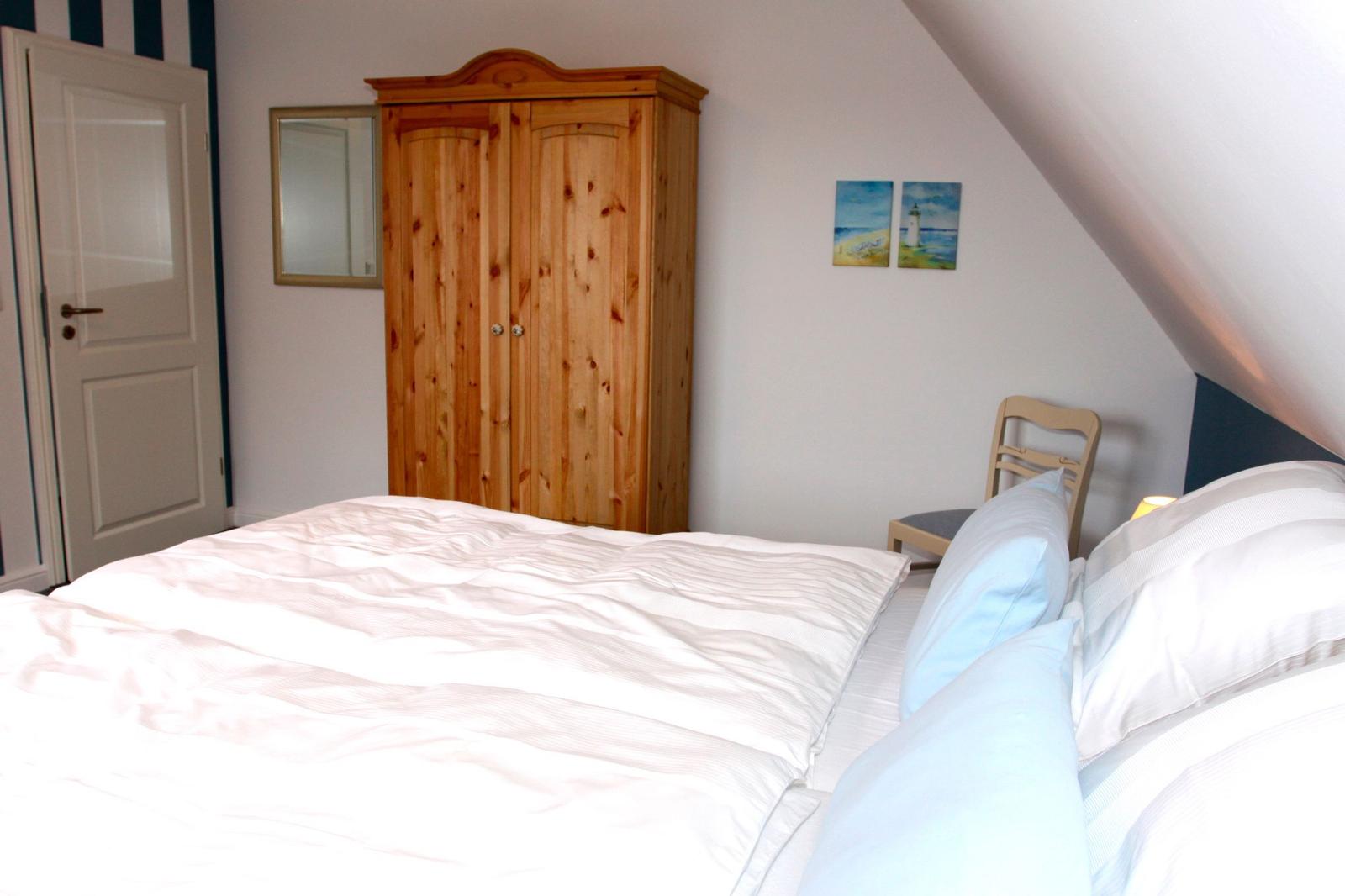 Schlafzimmer mit Dachschräge, Holzschrank und Bett mit weißen Laken.