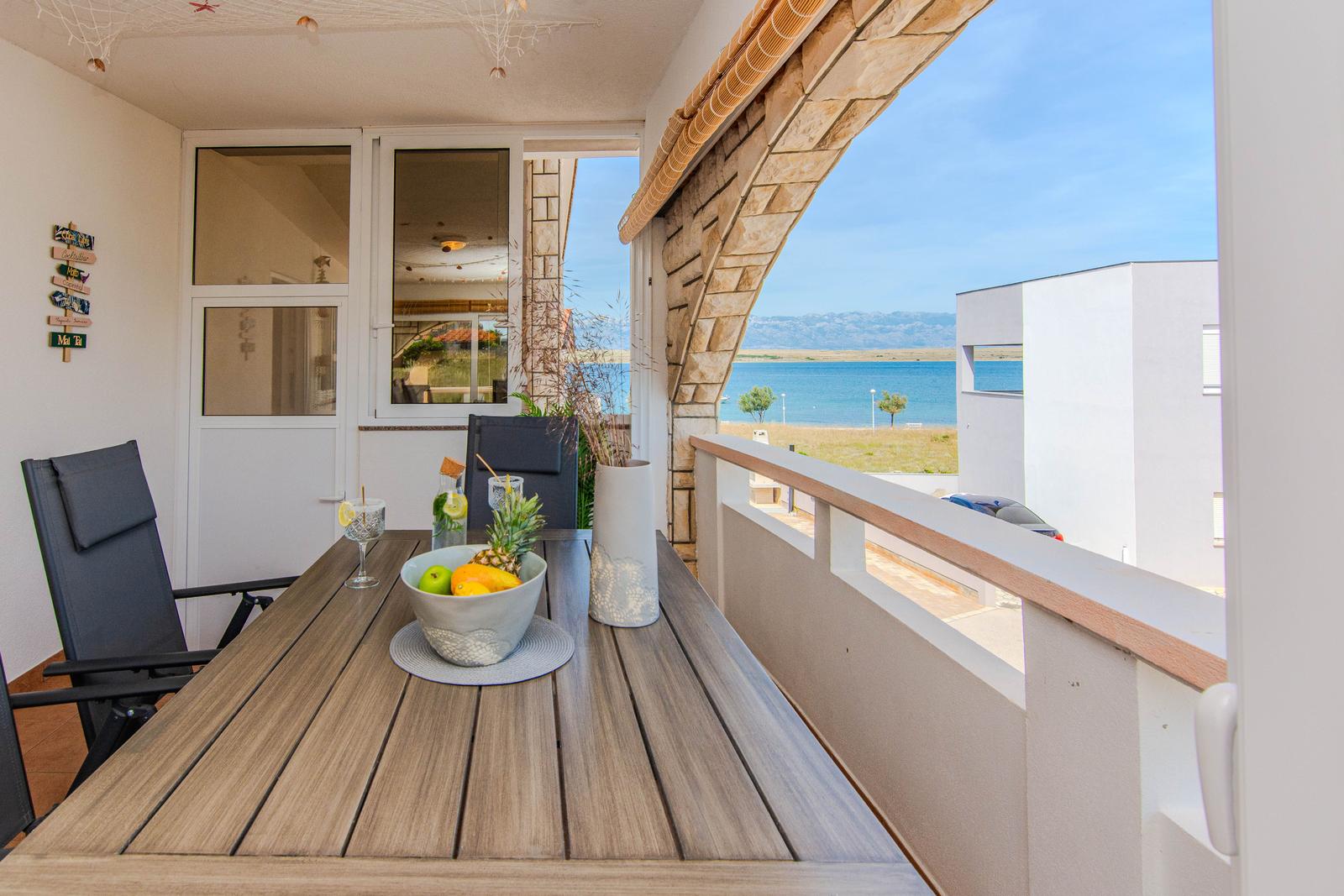 Terrasse mit Tisch, Stühlen und Meerblick. Tisch mit Obstschale und Getränken.
