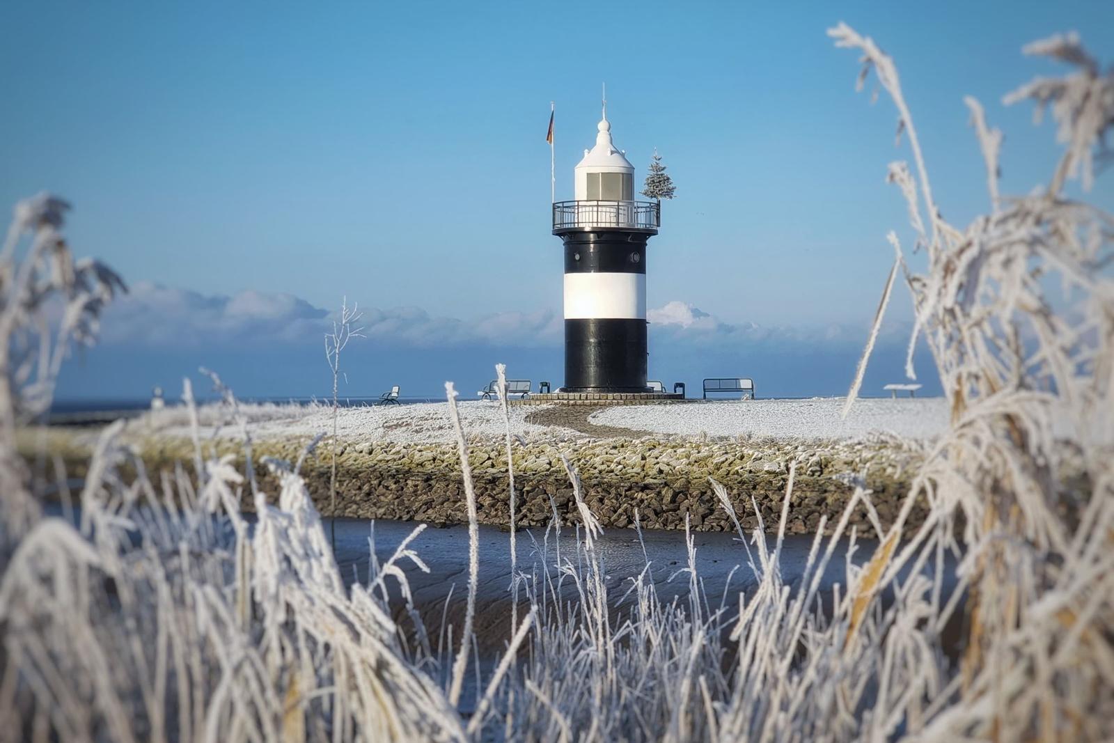 Frostiger Leuchtturm auf einem steinigen Ufer mit gefrorenem Gras im Vordergrund.
