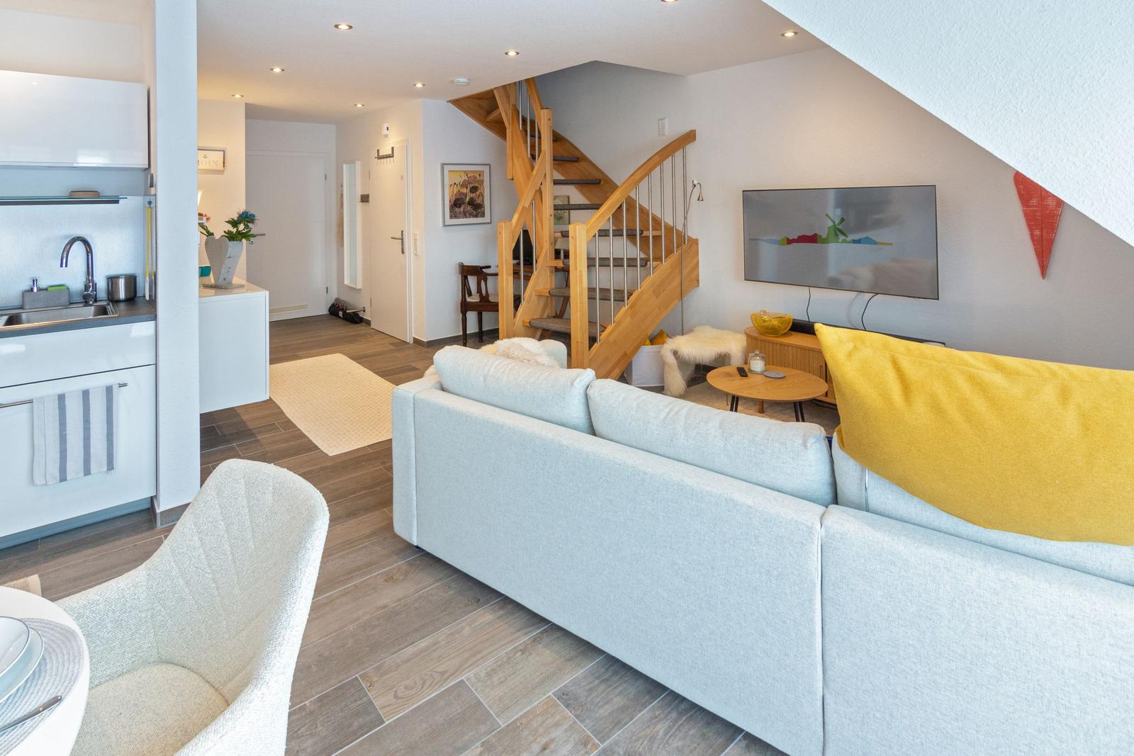Wohnzimmer mit Sofa, Fernseher und Holztreppe. Küche und Flur sind sichtbar.